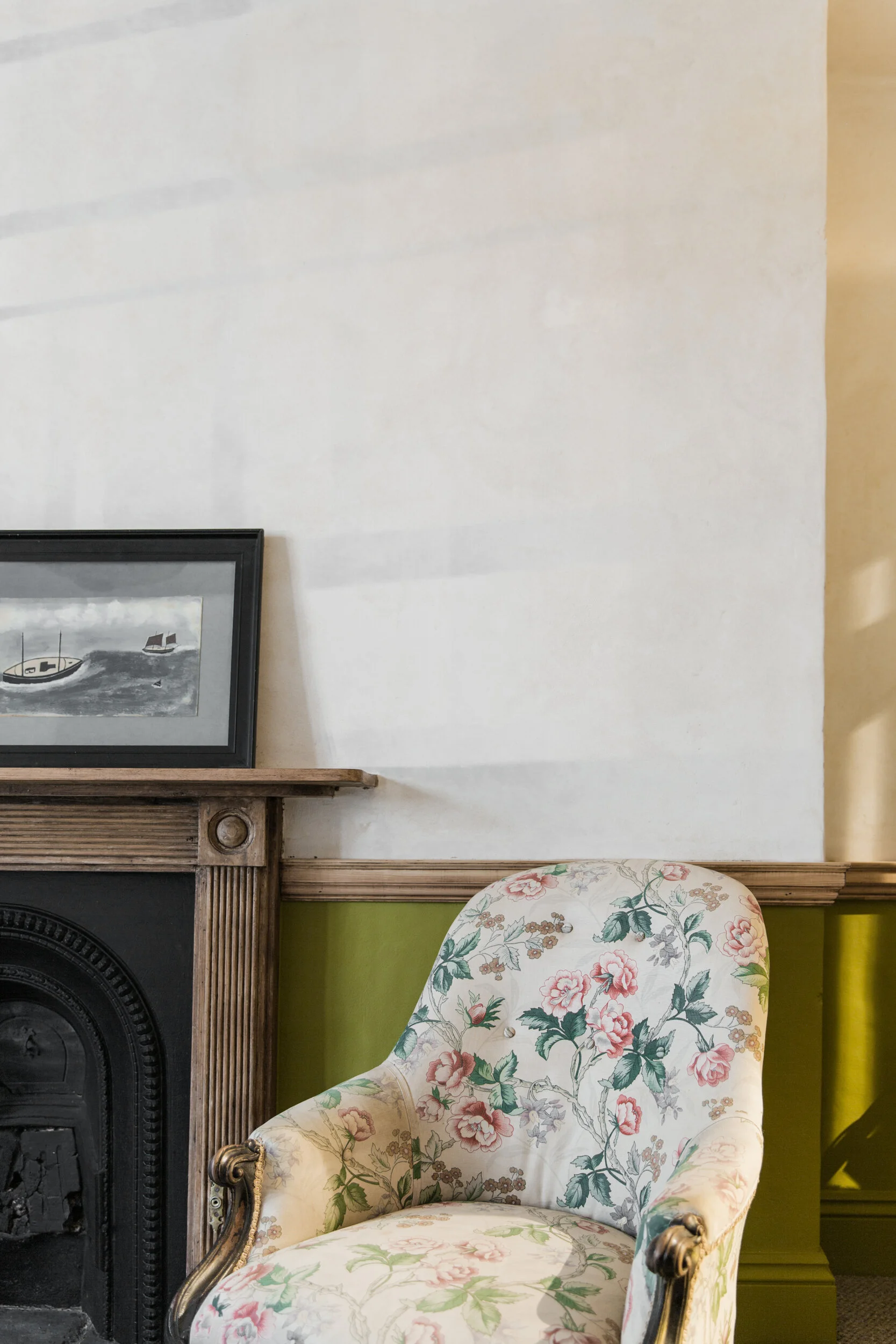 A corner of a room showing part of a black fireplace with a carved gold detail, a wooden mantel, a framed black and white picture of boats, a wall with a white and beige color, and a floral upholstered armchair with green, pink, and beige colors.