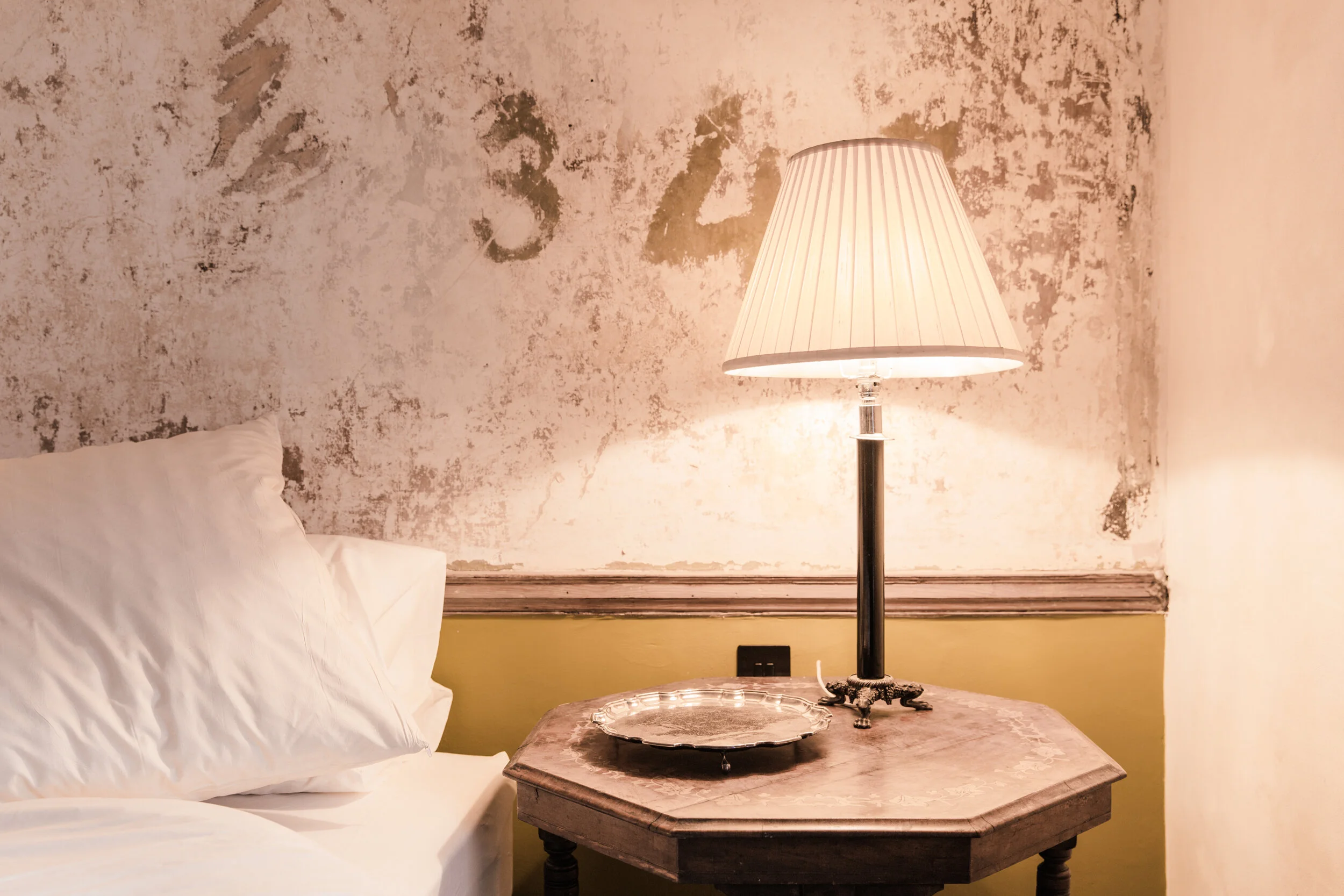 A bedside table with a glass tray, a vintage lamp with a pleated shade, against a distressed wall behind a bed with white bed linens.