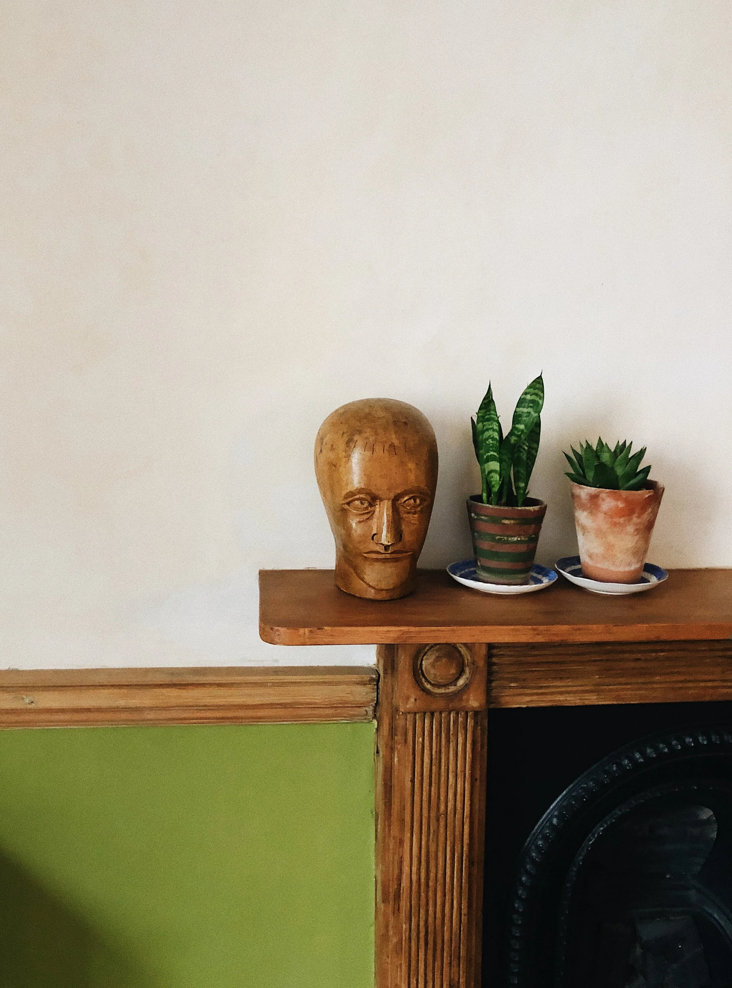 Wooden shelf with a carved wooden human head sculpture and two potted plants, one snake plant and one succulent, against a plain white wall.