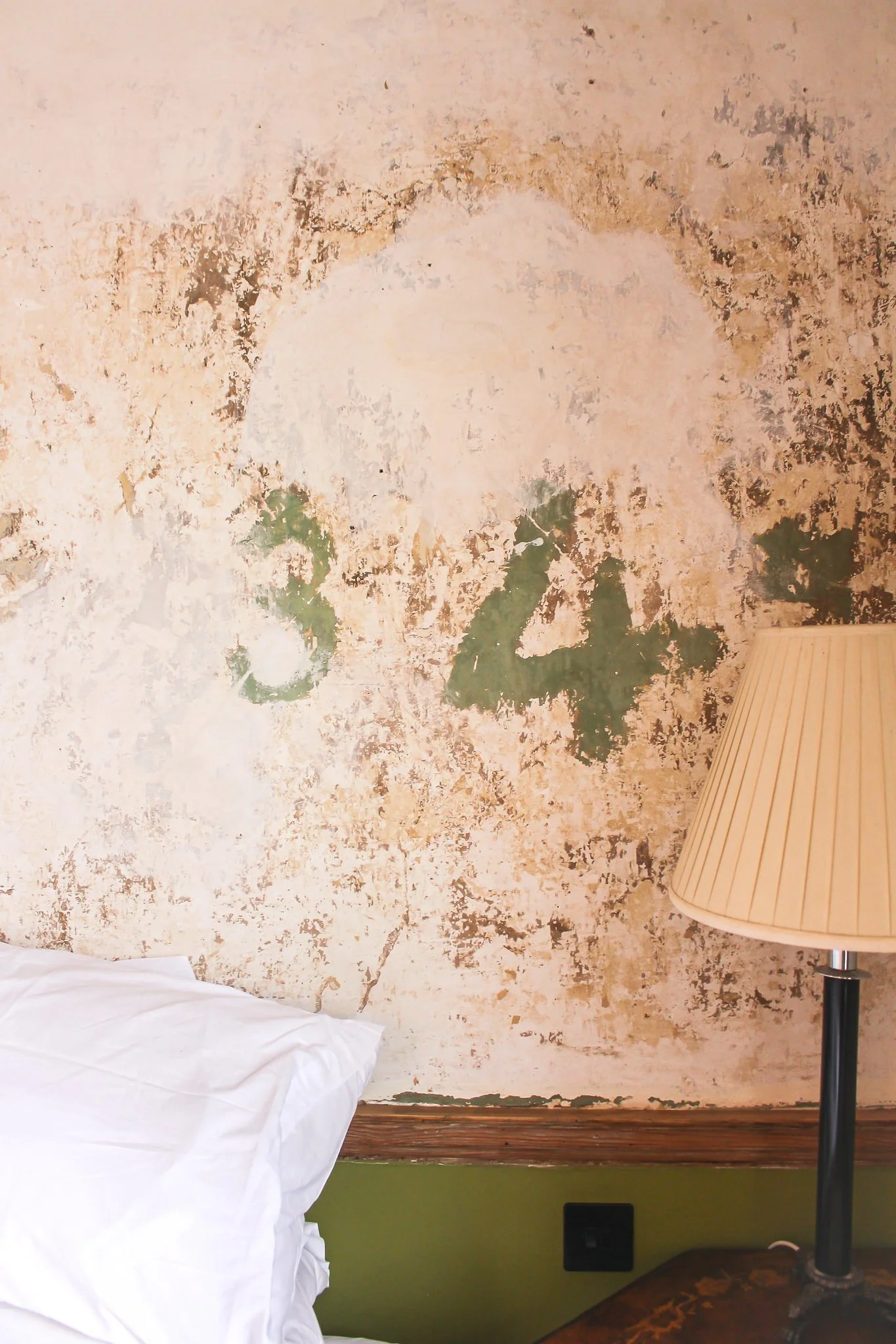 A wall with peeling paint and green underlayer, partially obscured by a bed with white linens and a floor lamp with a beige shade.