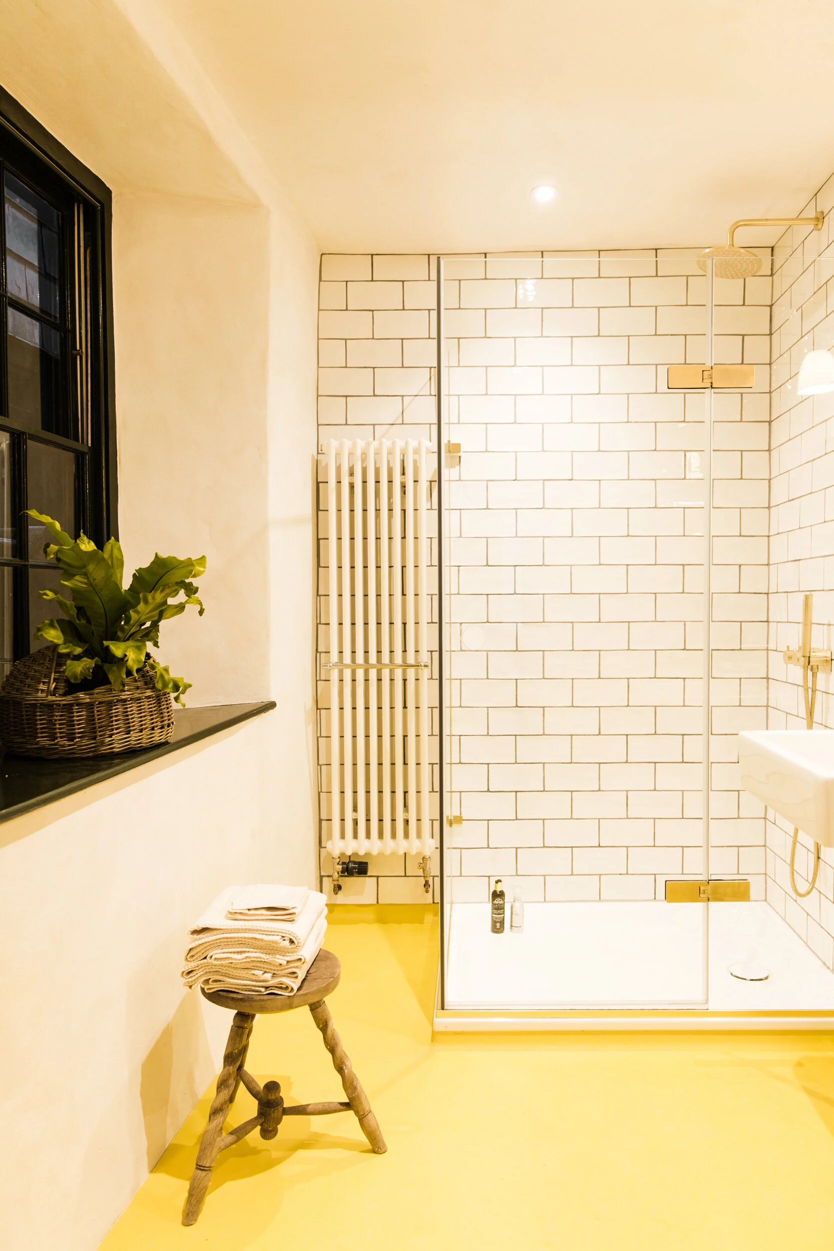 A bathroom with a yellow floor, white brick shower wall, showerhead, and a small wooden stool with folded towels, a potted plant on a ledge, and a white radiator.