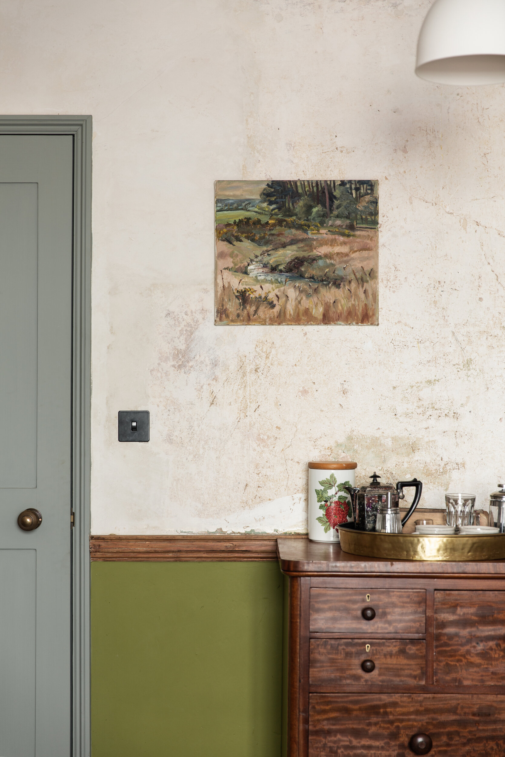 A cozy corner of a room with a textured beige wall and a green section at the bottom. There is a painting of a landscape with trees, a creek, and fields hanging on the wall. A wooden dresser with a dark finish holds a large white jar with a strawberr