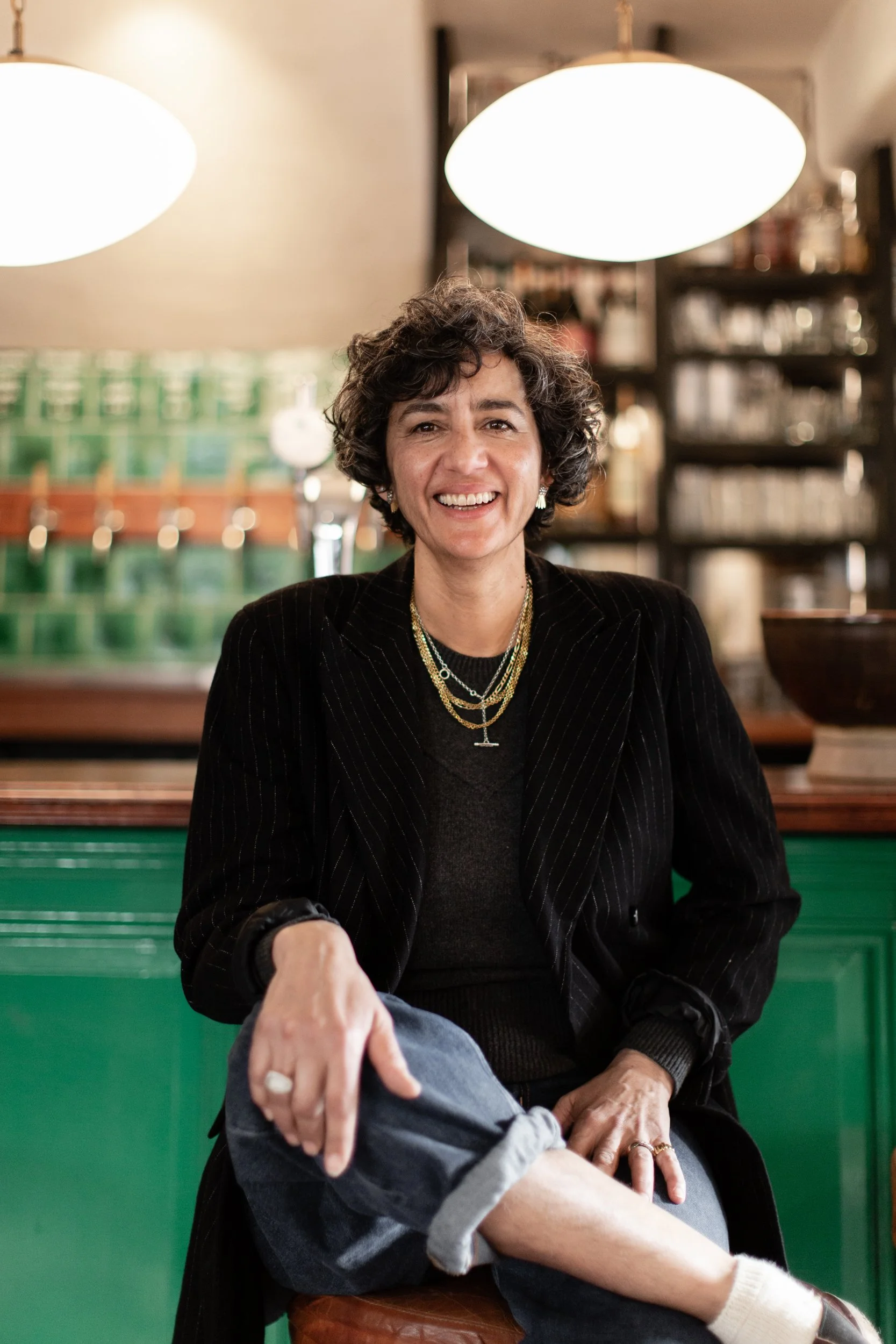 A woman with short curly hair smiling in a restaurant or cafe, with large hanging lights above her and shelves with bottles behind her.