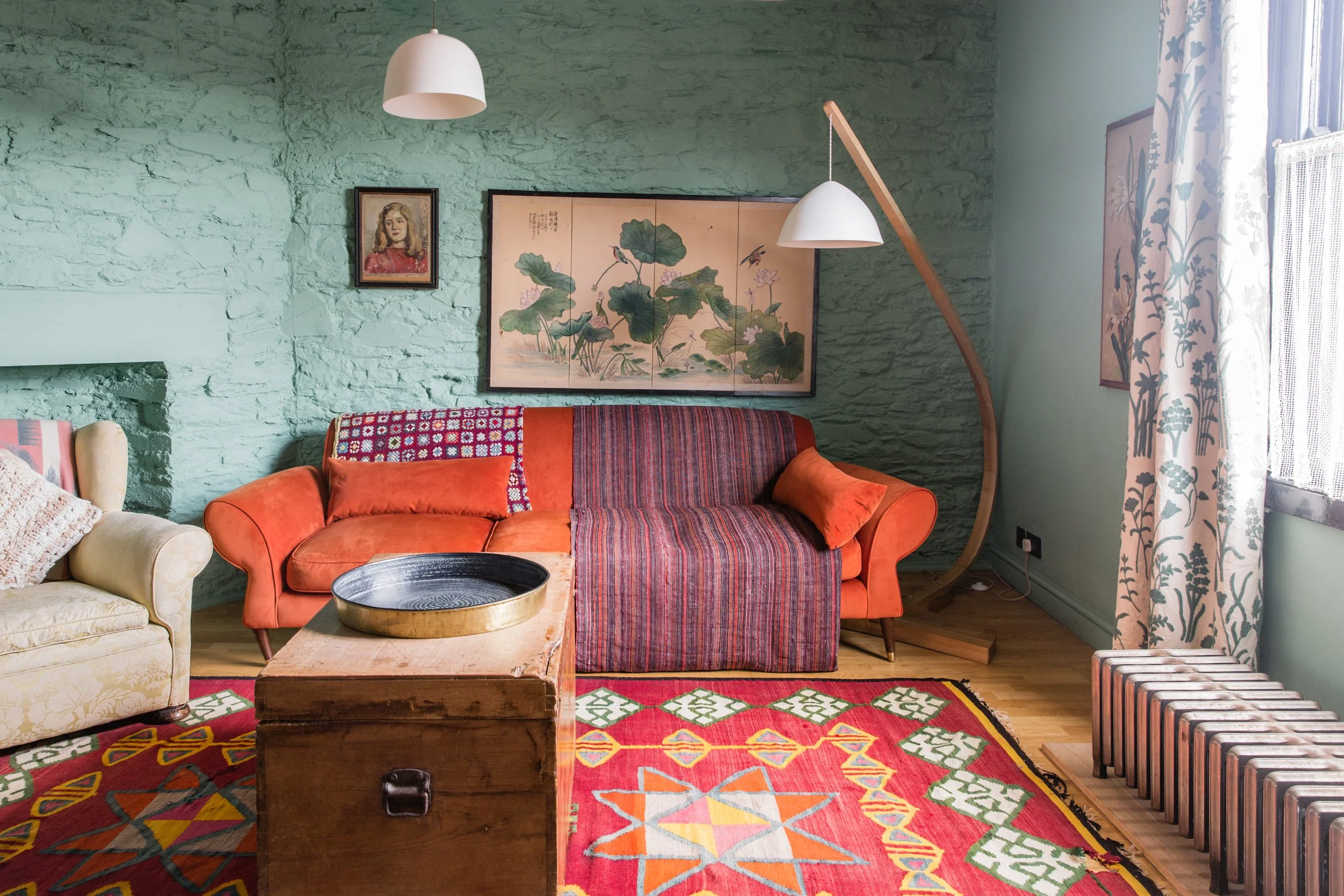 Living room with green textured walls, orange and cream upholstered sofas, a wooden coffee table with a round black tray, colorful patterned rugs, botanical artwork, a portrait, and a large window with floral curtains. Two modern white pendant lights hang from an arching wooden support.