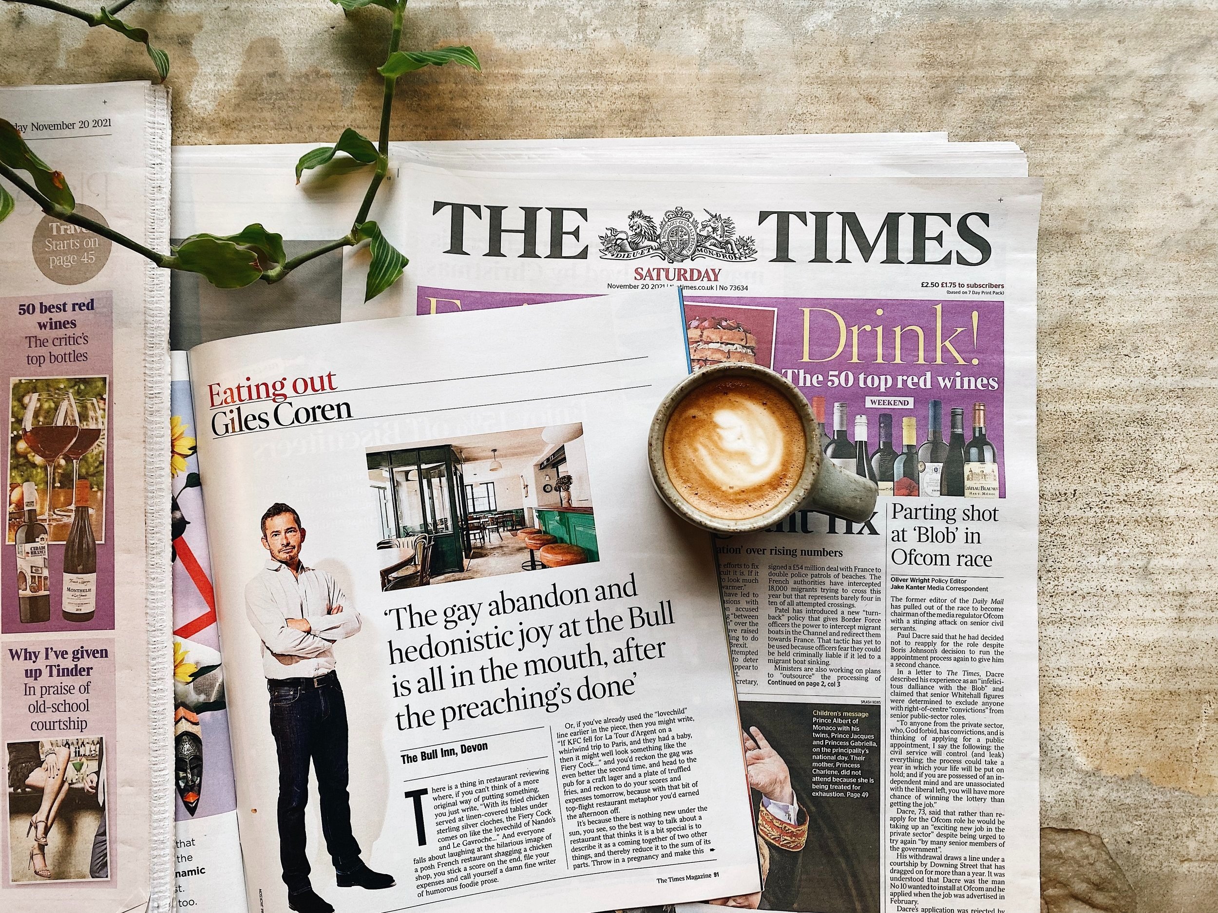 A newspaper titled The Times with a purple section, a cup of coffee with foam art, and a green plant on a textured beige surface.