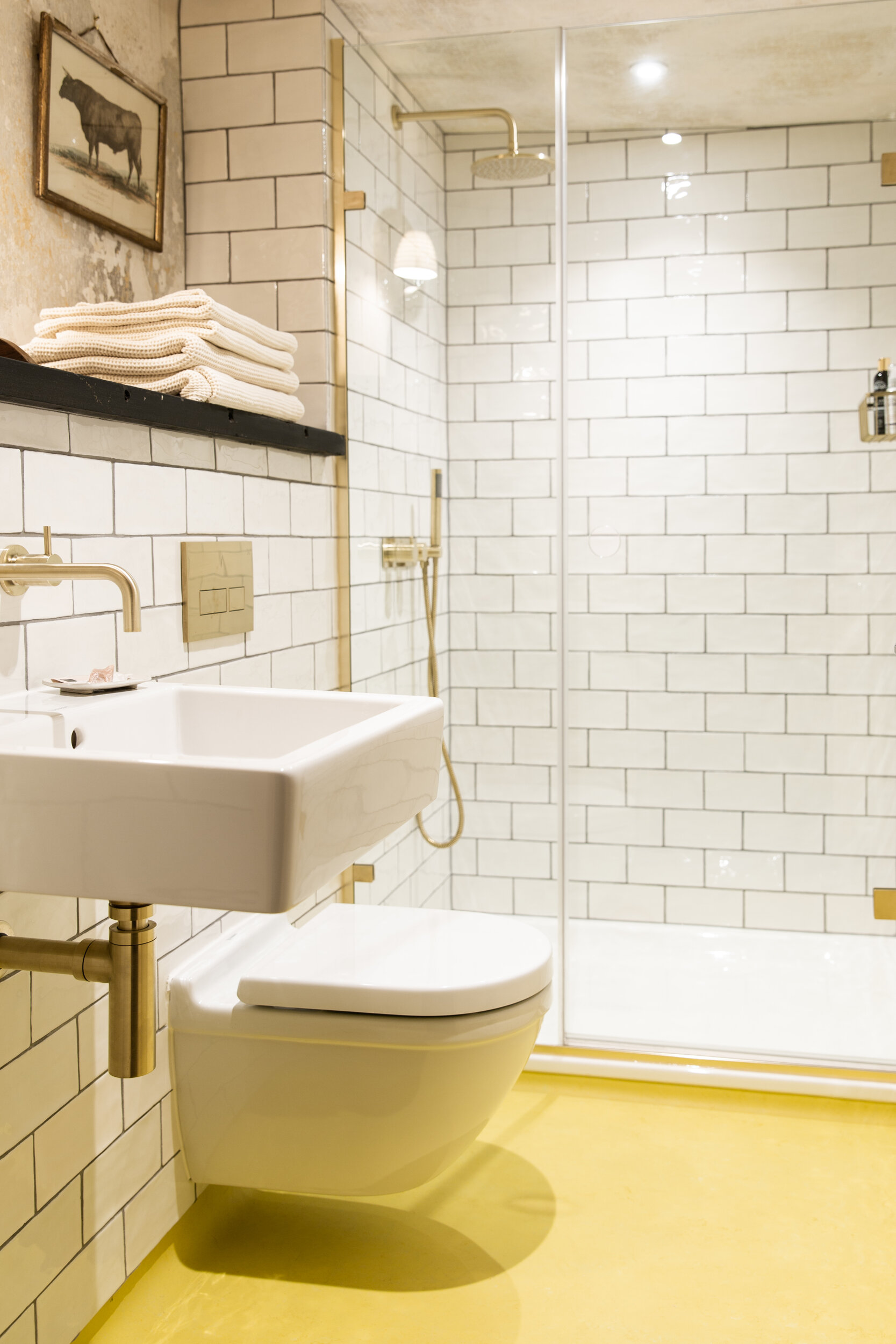 A modern bathroom with white subway tile walls, a white toilet, a white wall-mounted sink, a gold showerhead, and folded white towels on a dark shelf. There is a small framed elephant artwork on the wall and a yellow floor.