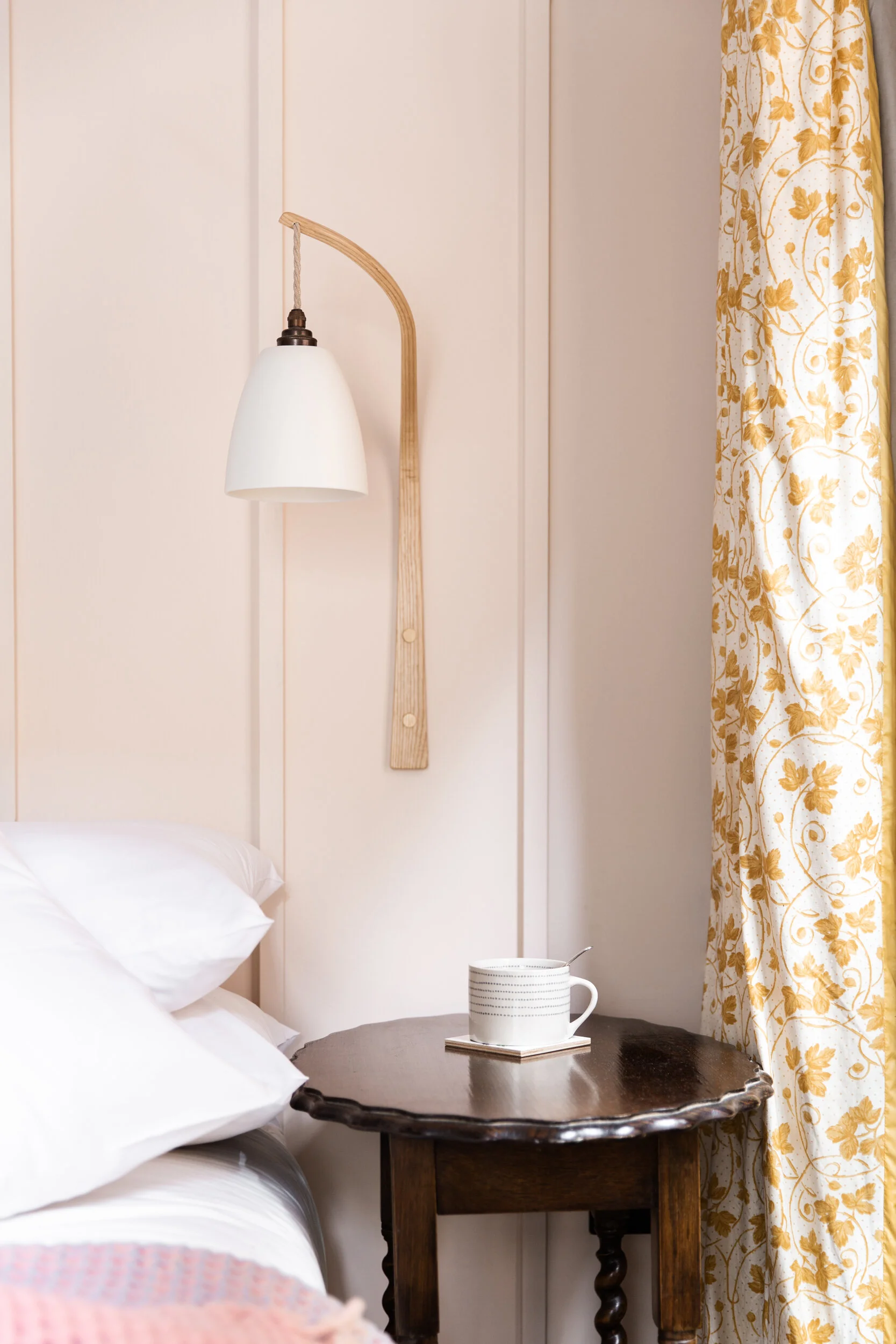 Close-up of a bedroom nightstand with a white mug on a book, next to a bed with white pillows, and a wall-mounted lamp with a wooden arm and a white conical shade. Yellow floral curtains are partly visible on the right side.