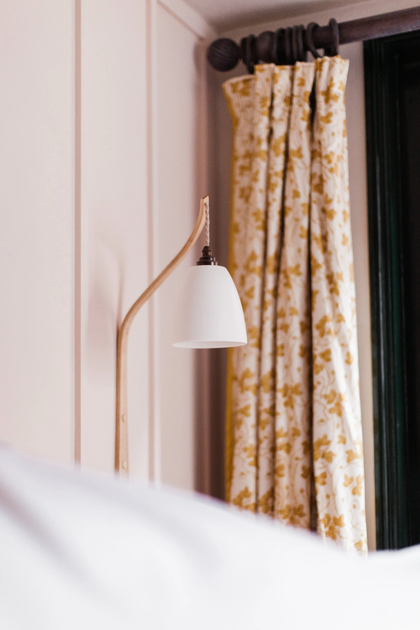 Close-up of a wall-mounted white lamp with a curved wooden arm, in front of a window with floral yellow and white curtains.