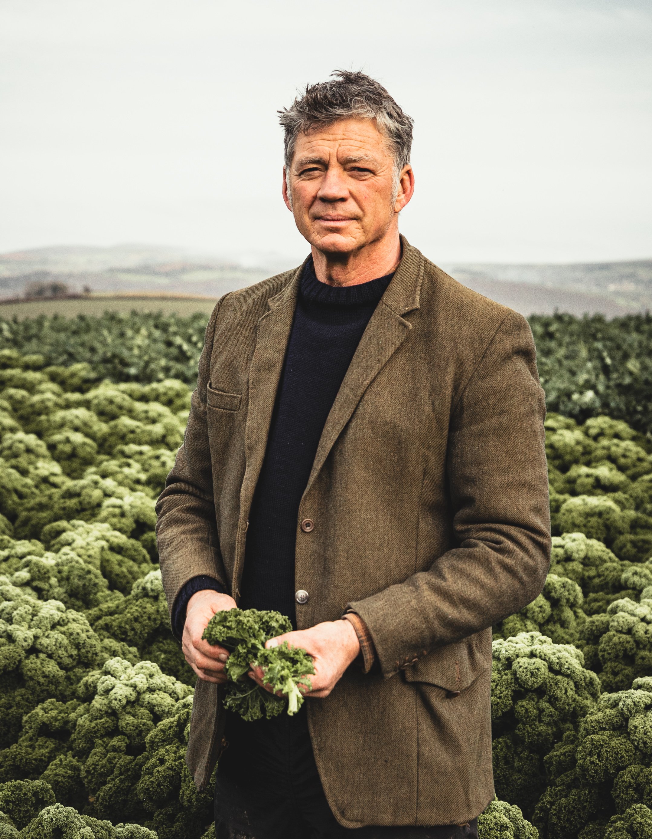 A man standing in a lush green field holding some greenery, wearing a brown blazer and a black shirt, with rolling hills in the background.