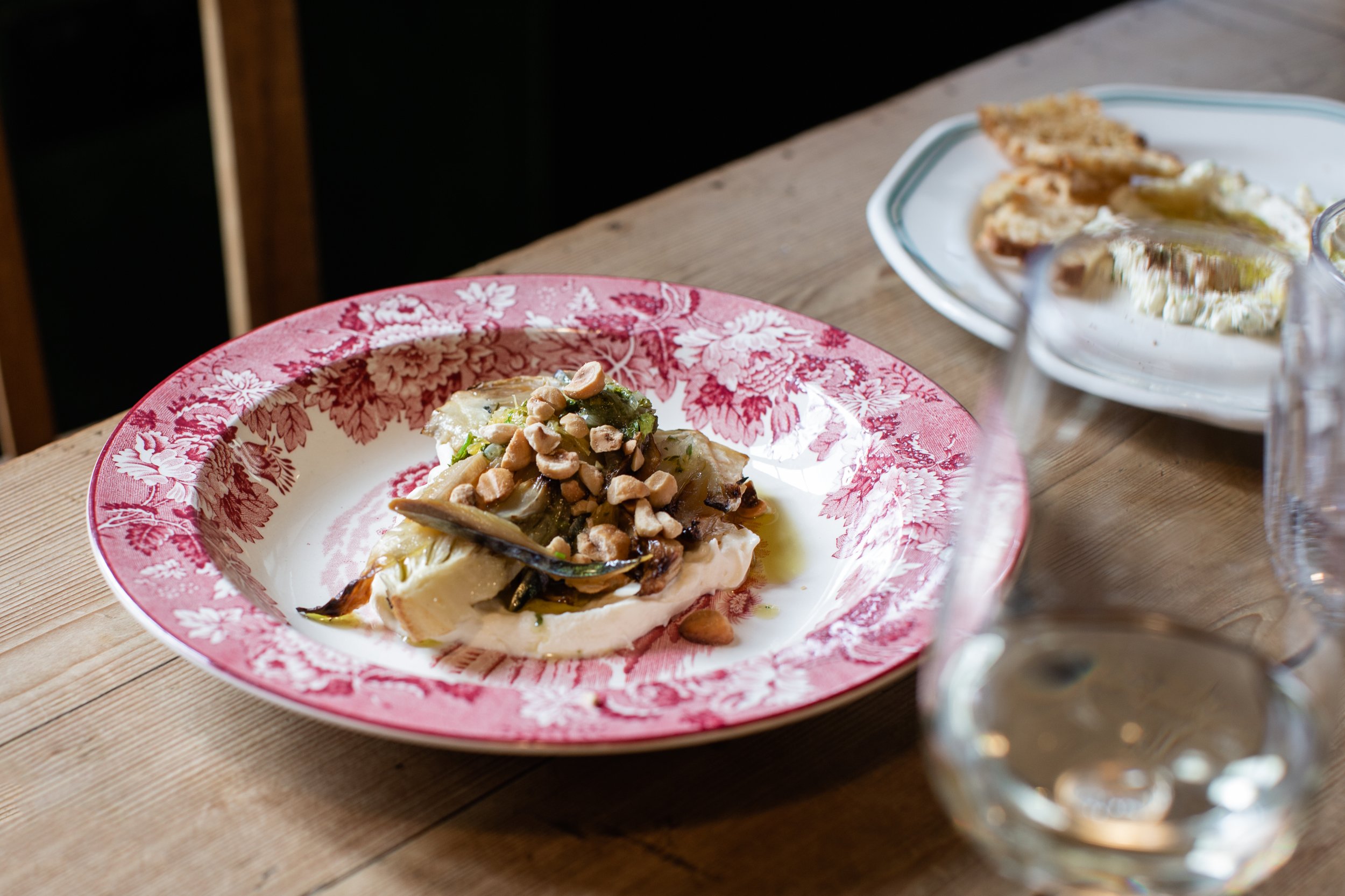 A plate of food with a piece of fish topped with chopped nuts and sauce, served on a red and white floral plate on a wooden table, with another plate of food and a glass in the background.