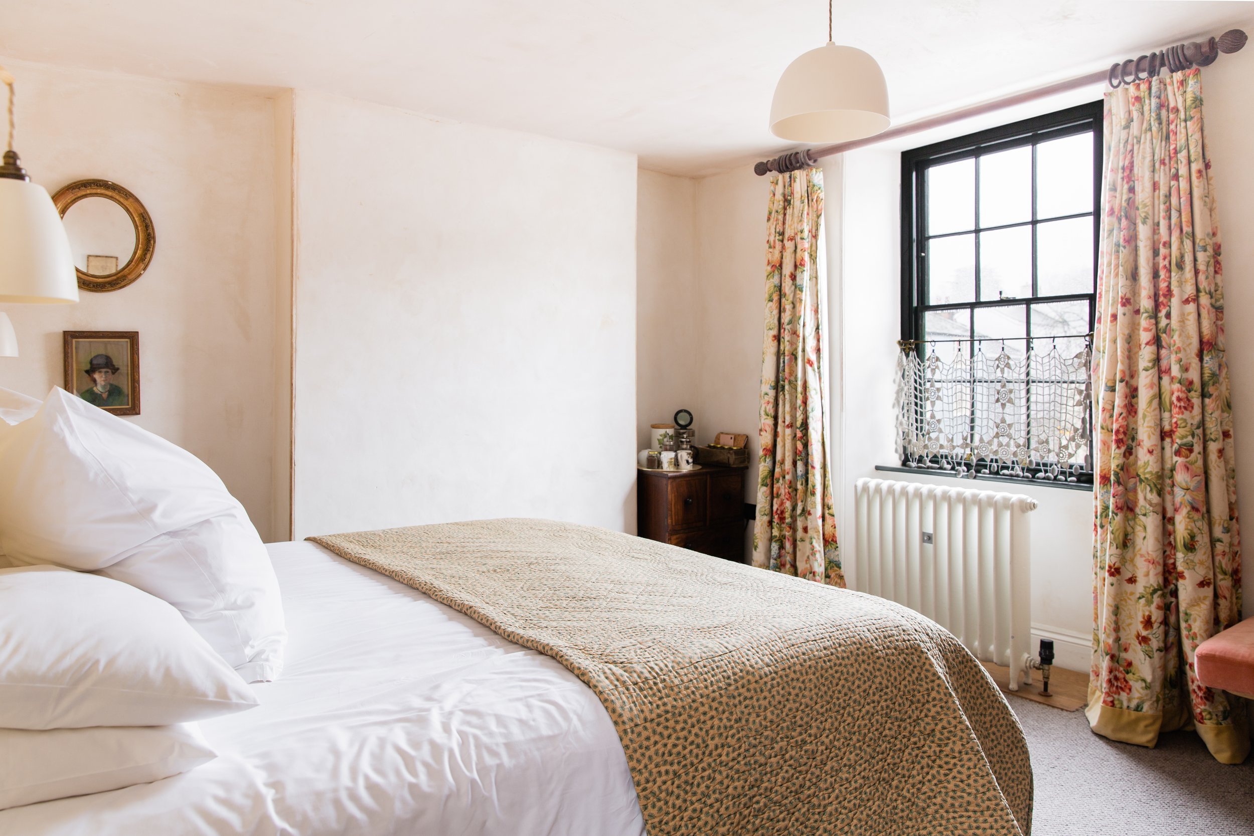 A cozy bedroom features a bed with white pillows and a beige patterned blanket, floral curtains by a large black-framed window, a small wooden side table with various items, and a radiator underneath the window. The room has vintage-style artwork and a hanging white pendant light.
