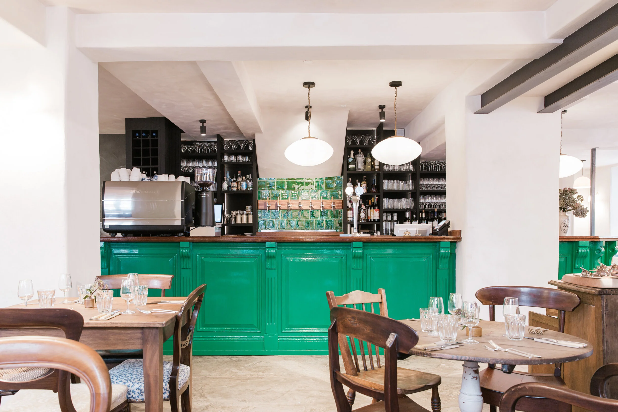 Interior view of a restaurant or cafe with a bar area, wooden tables, chairs, and decor including glasses, bottles, and plants, with hanging pendant lights and a green-paneled counter.