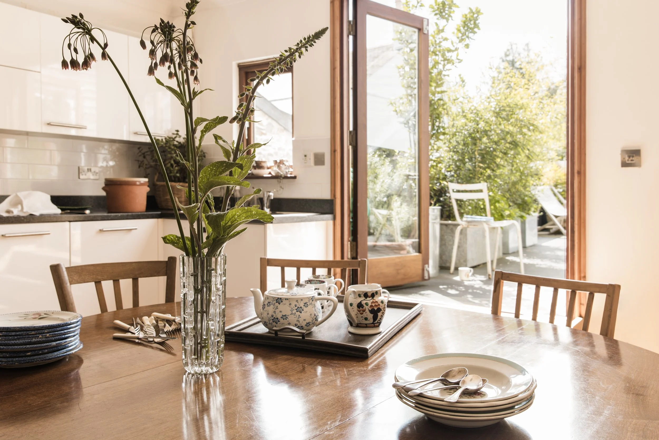 A dining table set with plates, teapot, cups, and cutlery, with a glass vase holding green plants, in a room with open wooden doors leading to an outdoor patio with chairs and lush greenery.