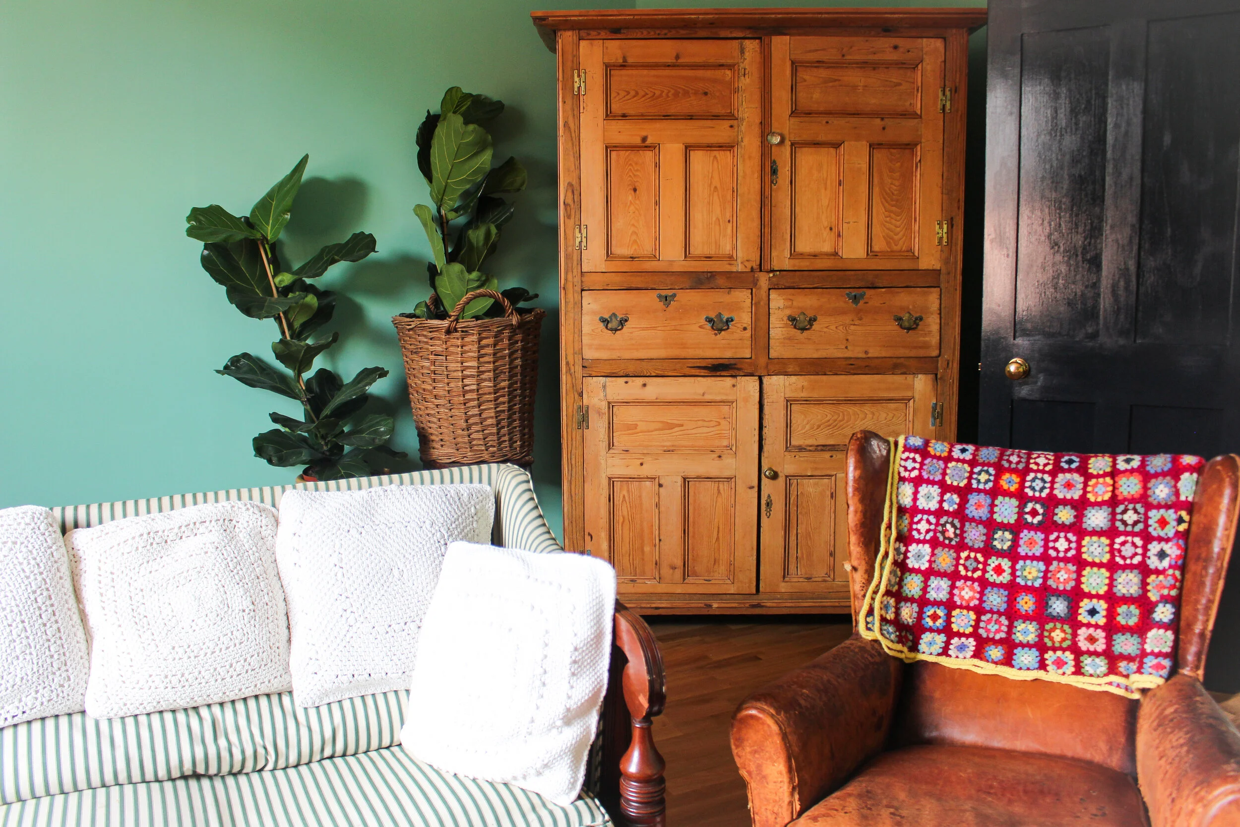 Living room with a green wall, a tall potted fiddle leaf fig plant in a woven basket, a white crocheted throw on a green and white striped sofa, a wooden cabinet with brass hinges, a black door, and a red wooden chair with a crocheted granny square b
