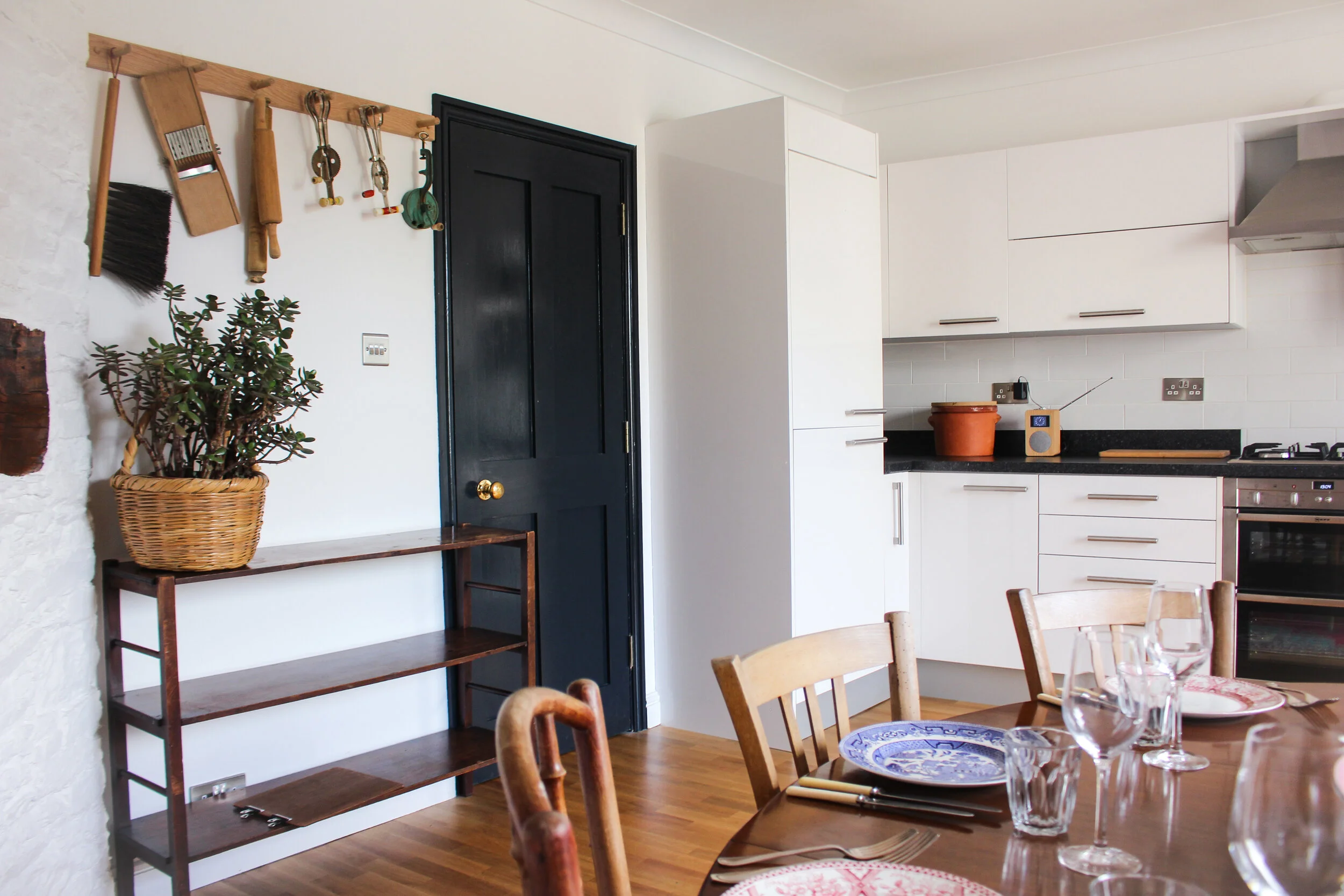 A kitchen with white cabinets, a black door, and a wooden table set for dining with wine glasses and plates. A wooden shelf with a potted plant is on the left wall. Kitchen utensils are hanging on the wall near the door.