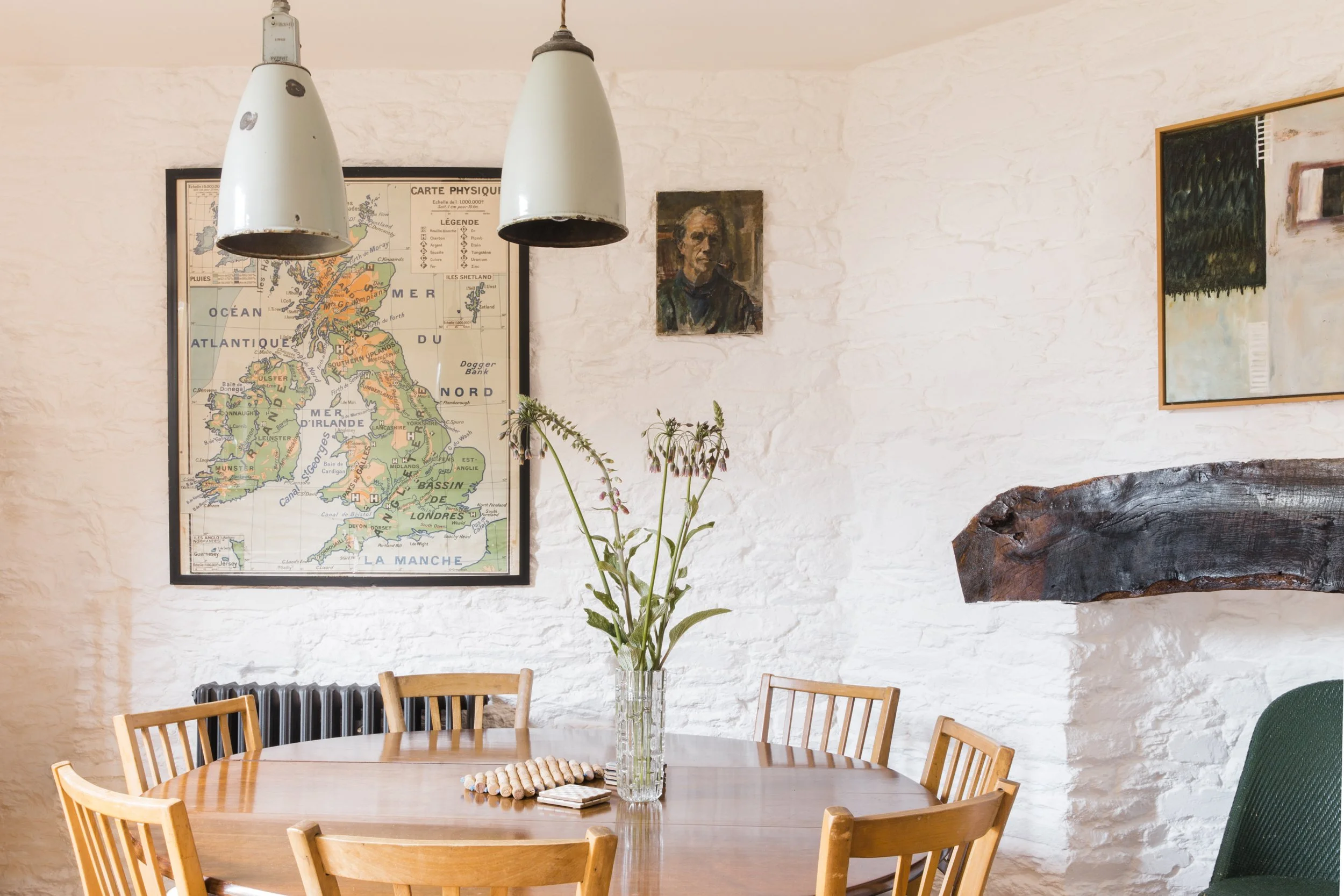 A dining room with a round wooden table surrounded by wooden chairs, a tall glass vase with green plants, and wall art including a map of Scotland, a portrait of a man with a cigarette, and an abstract painting, against a white textured brick wall.