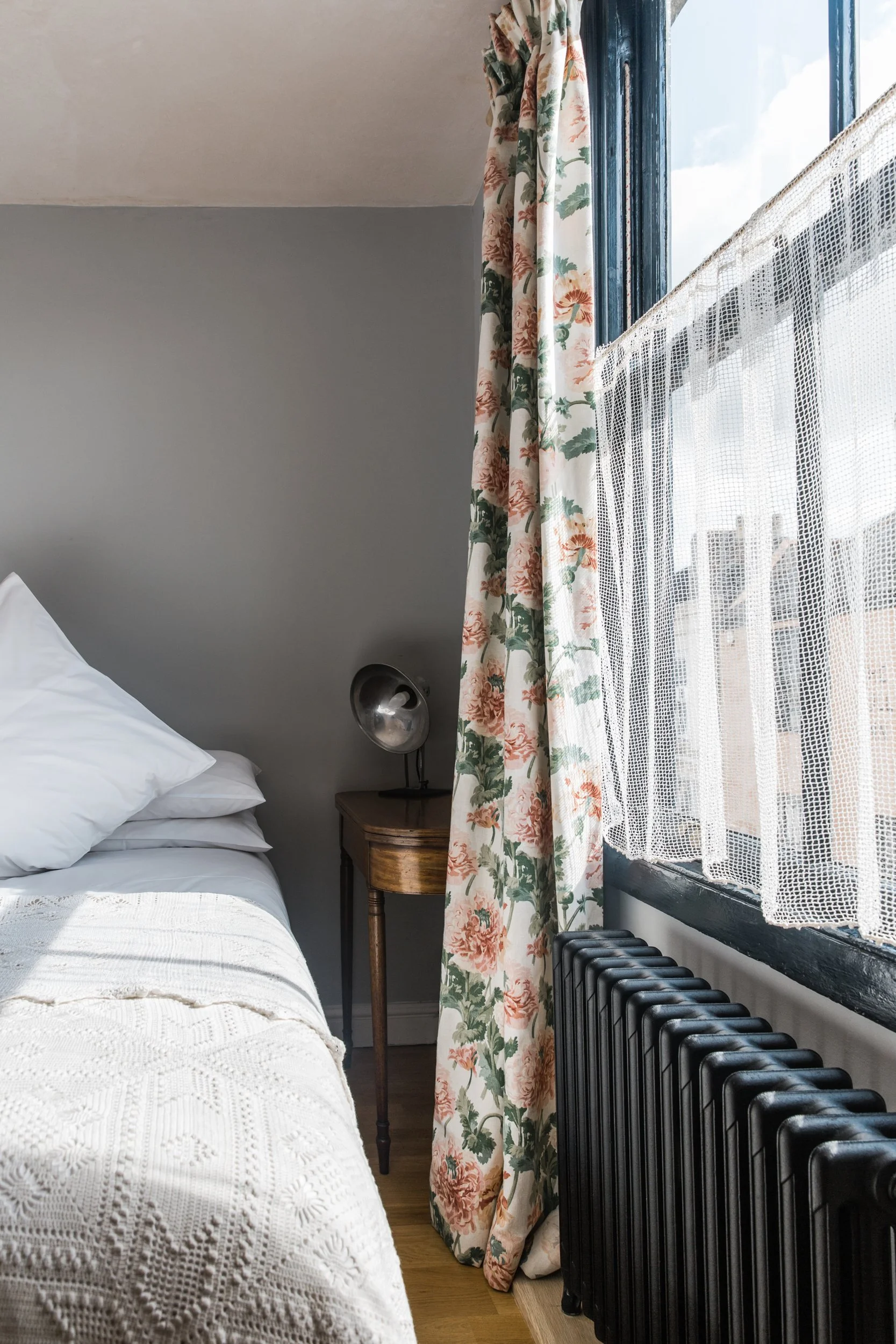 A cozy bedroom with a white bed, a wooden nightstand, and a vintage black radiator beneath a large window. The window has sheer white curtains and floral drapes, allowing natural light to brighten the room, and a metallic wall lamp is mounted beside 