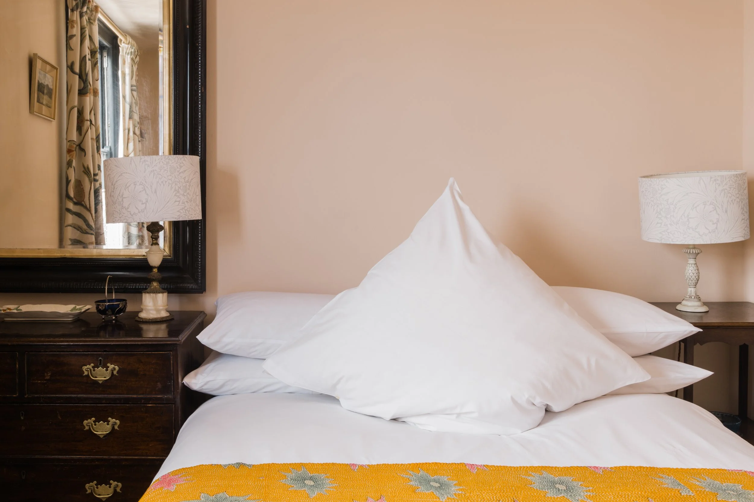 A neatly made bed with white sheets and pillows, flanked by nightstands with lamps, a full-length mirror, and floral curtains in a warmly lit bedroom.