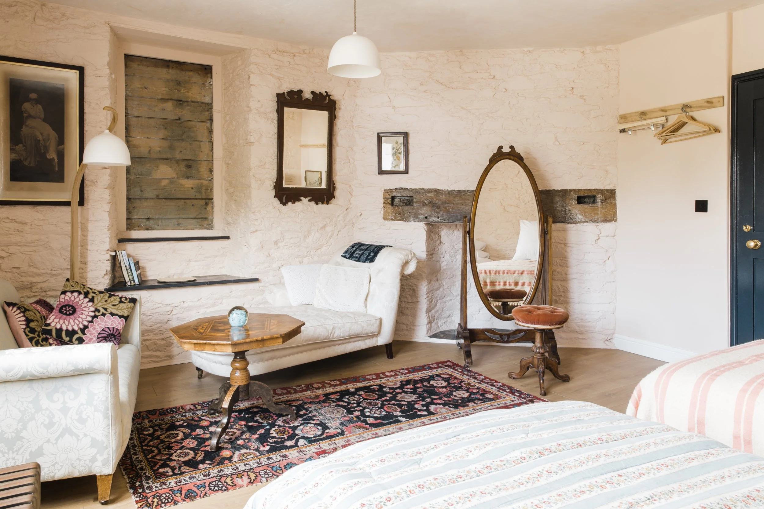 Interior of a cozy bedroom with vintage furniture, including a white upholstered armchair with floral cushions, a wooden side table, a standing mirror with a wooden frame, and a bed with a striped bedspread. The room features textured white walls, fr