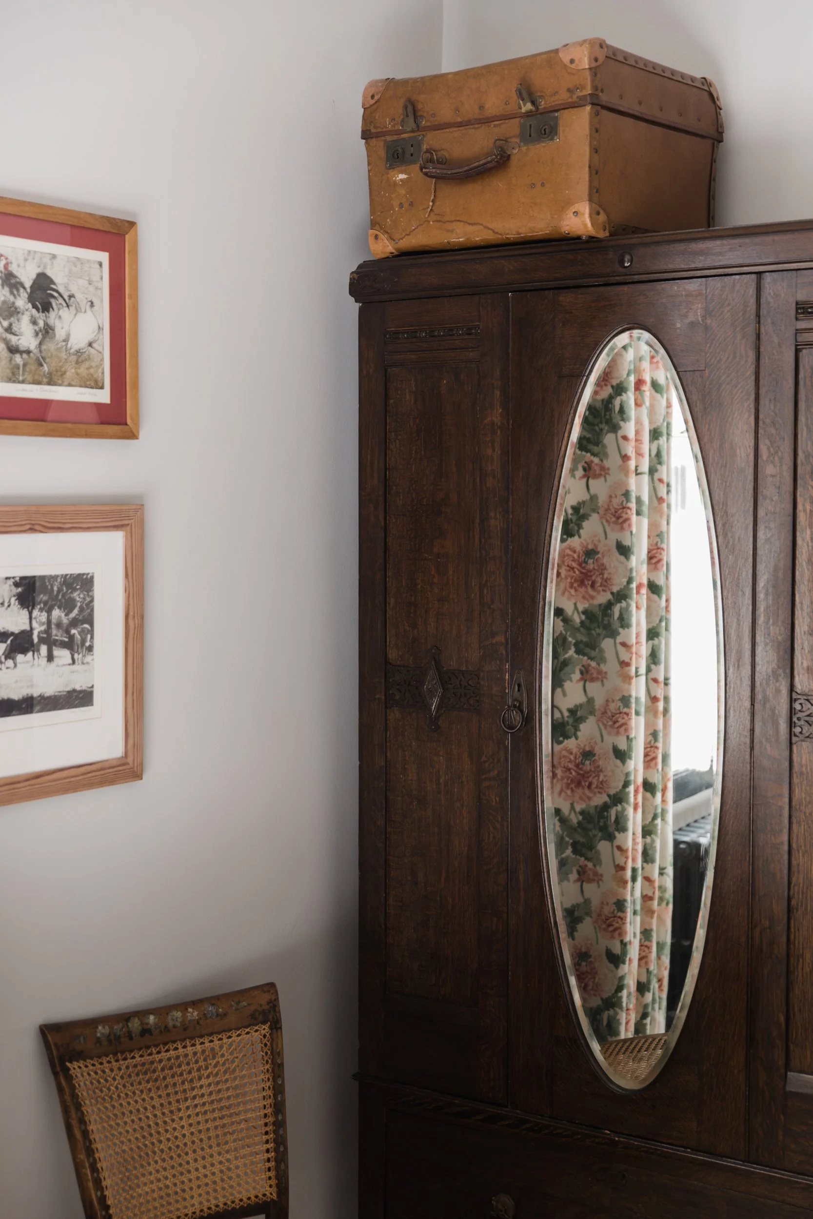 An antique wooden wardrobe with a rounded mirror hanging on the door, floral curtains reflected in the mirror, a vintage leather suitcase on top of the wardrobe, framed pictures on the white wall, and a wooden chair with a woven cane seat next to the