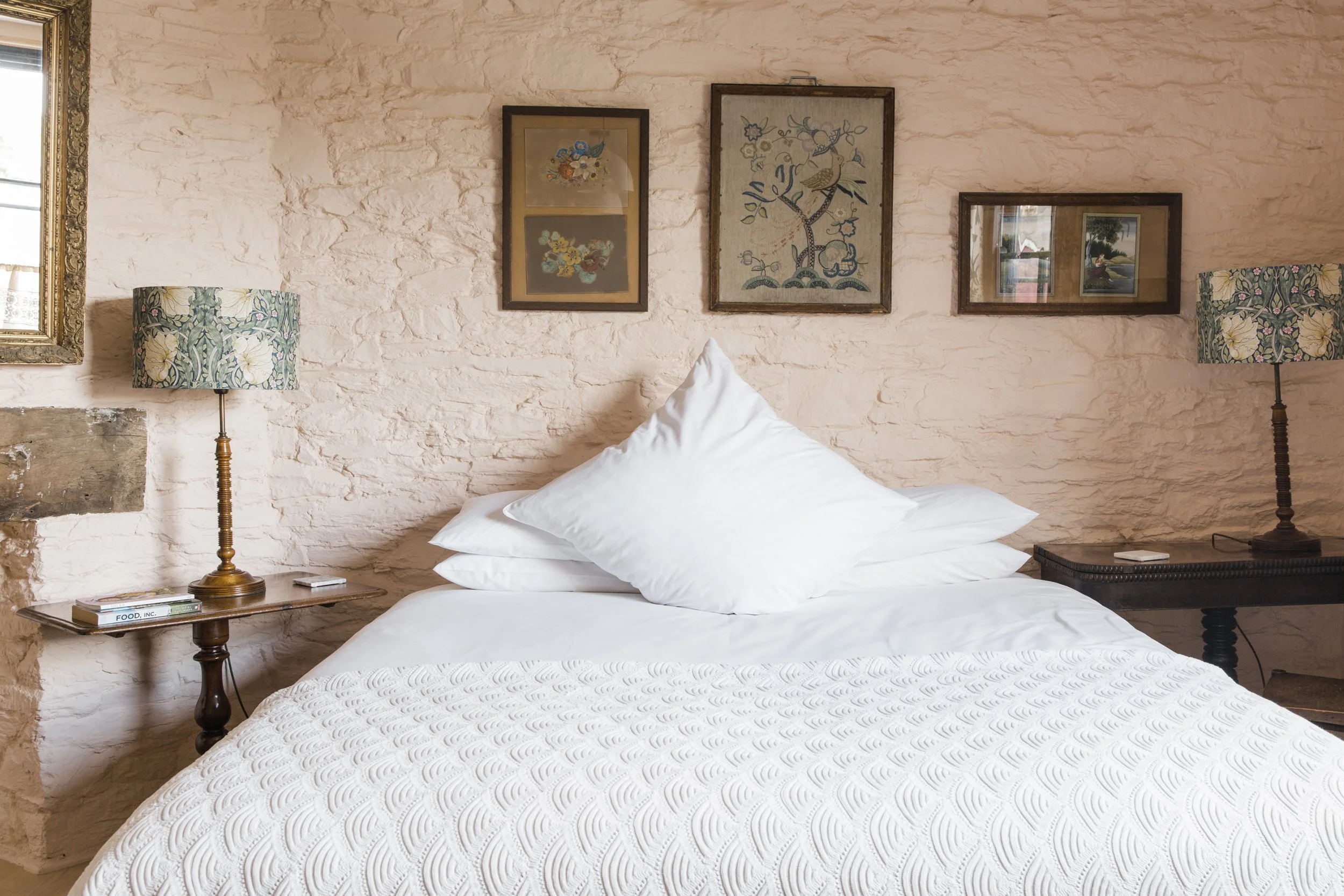 A bedroom with a bed covered in a white textured quilt, two white pillows, two wooden nightstands with table lamps featuring floral patterned shades, and framed artwork hanging on a textured stone wall behind the bed.