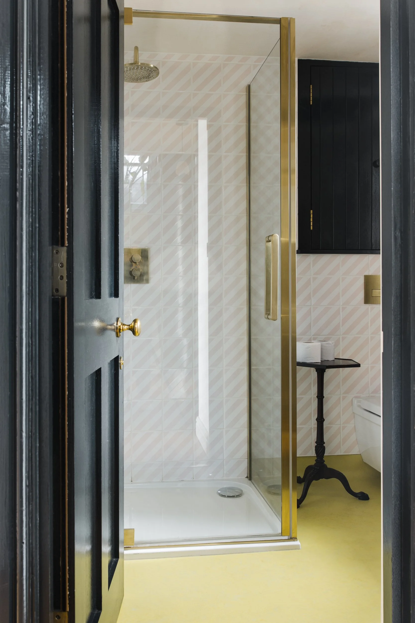 A bathroom with a glass shower door, a gold showerhead, and gold door handle. Cream tiled walls with a diagonal stripe pattern, a black cabinet, and a small black table holding toilet paper rolls. The floor is yellow, and part of a toilet is visible in the corner.