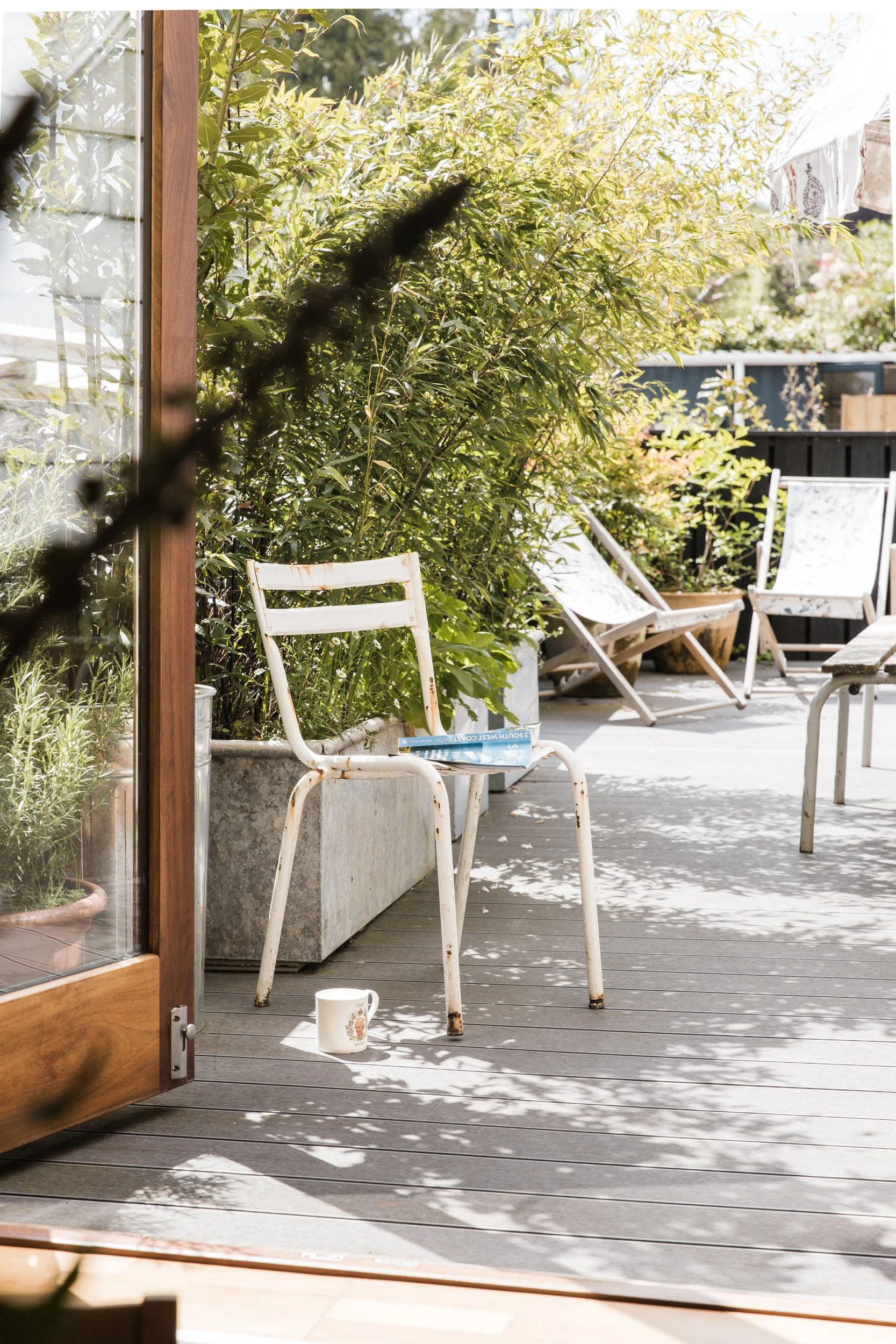 A sunlit outdoor patio with wooden flooring, white metal chairs, potted plants, and lush green bushes in the background.