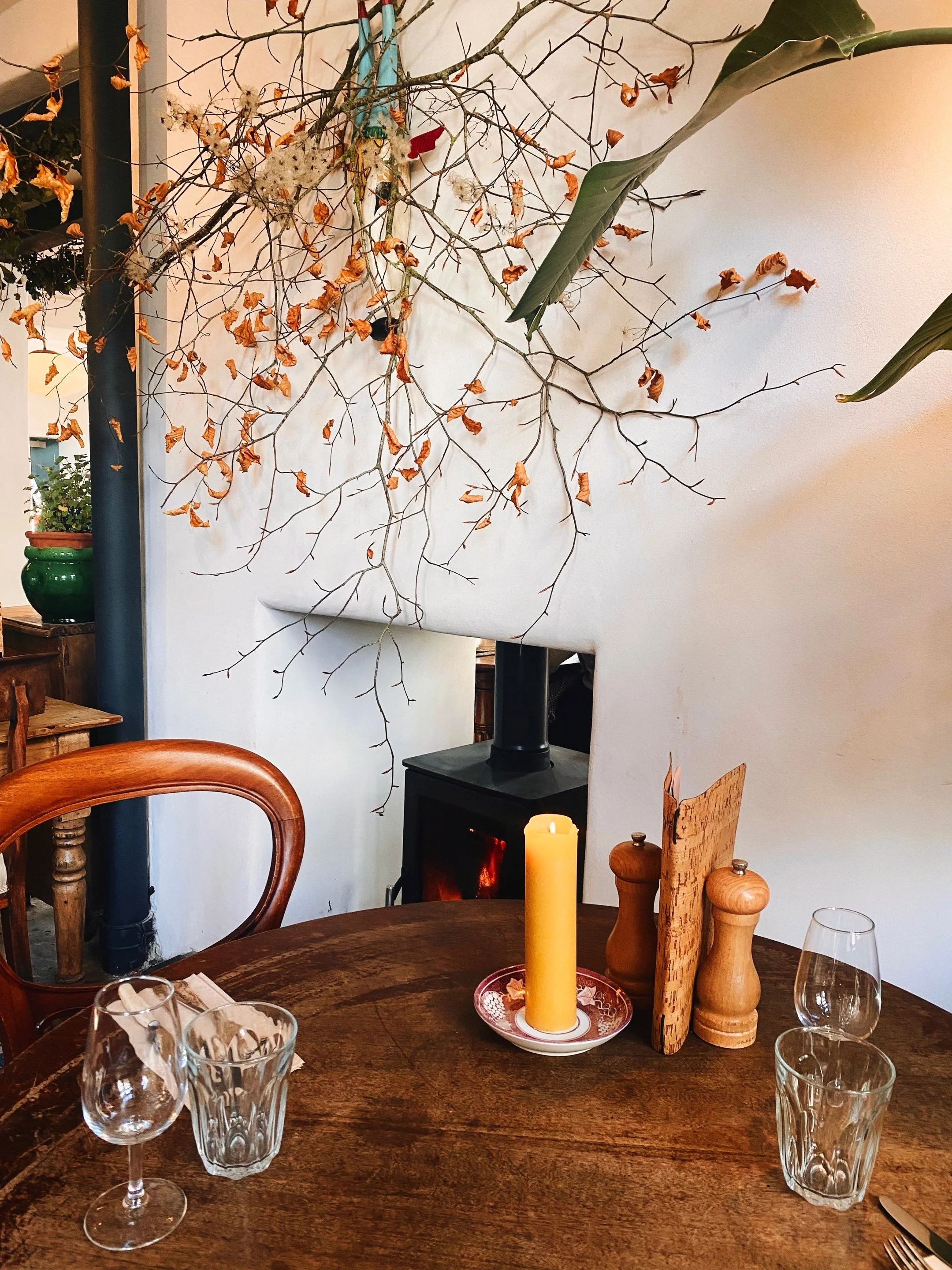 A wooden table set with four glasses and a candle on a decorative plate, surrounded by wooden salt and pepper shakers and a glass of water. In the background is a decorative wall with dried branches and leaves, and kitchen items including a green pot