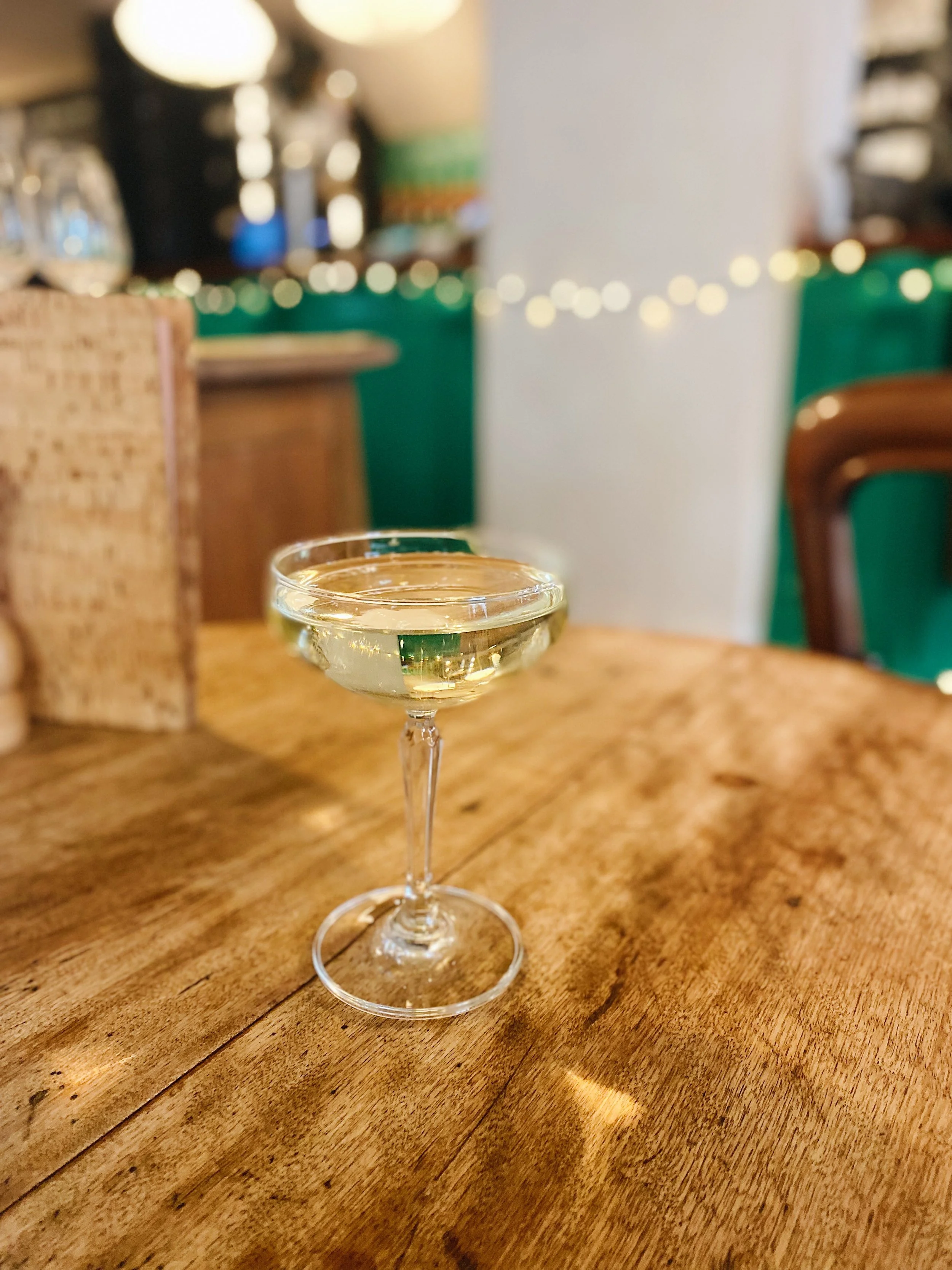 A glass of white wine on a wooden table in a cozy, decorated restaurant or bar with warm lighting and string lights in the background.