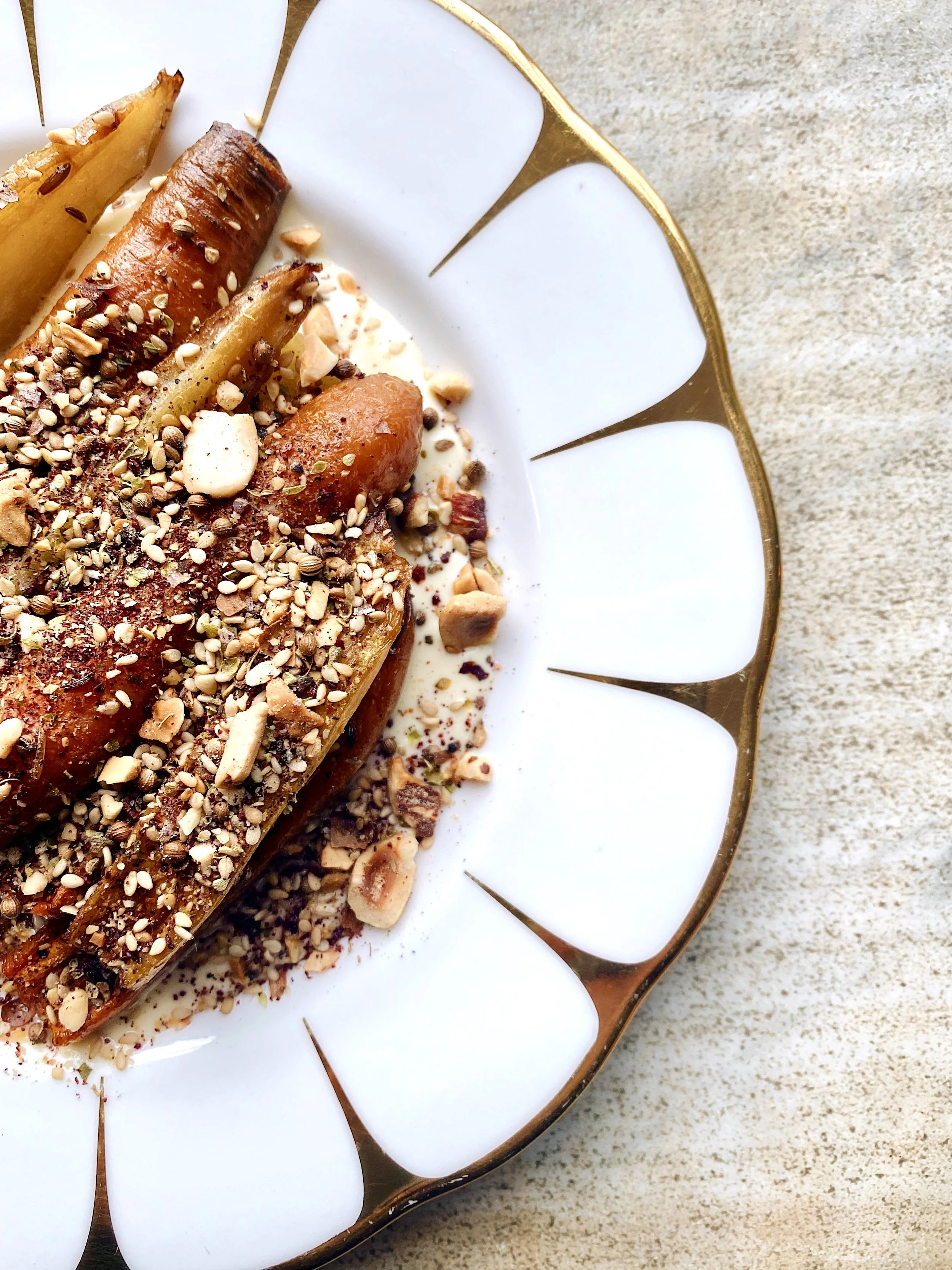 A white decorated plate with a gold rim holding a dessert topped with nuts, seeds, and spices, set on a light-colored surface.