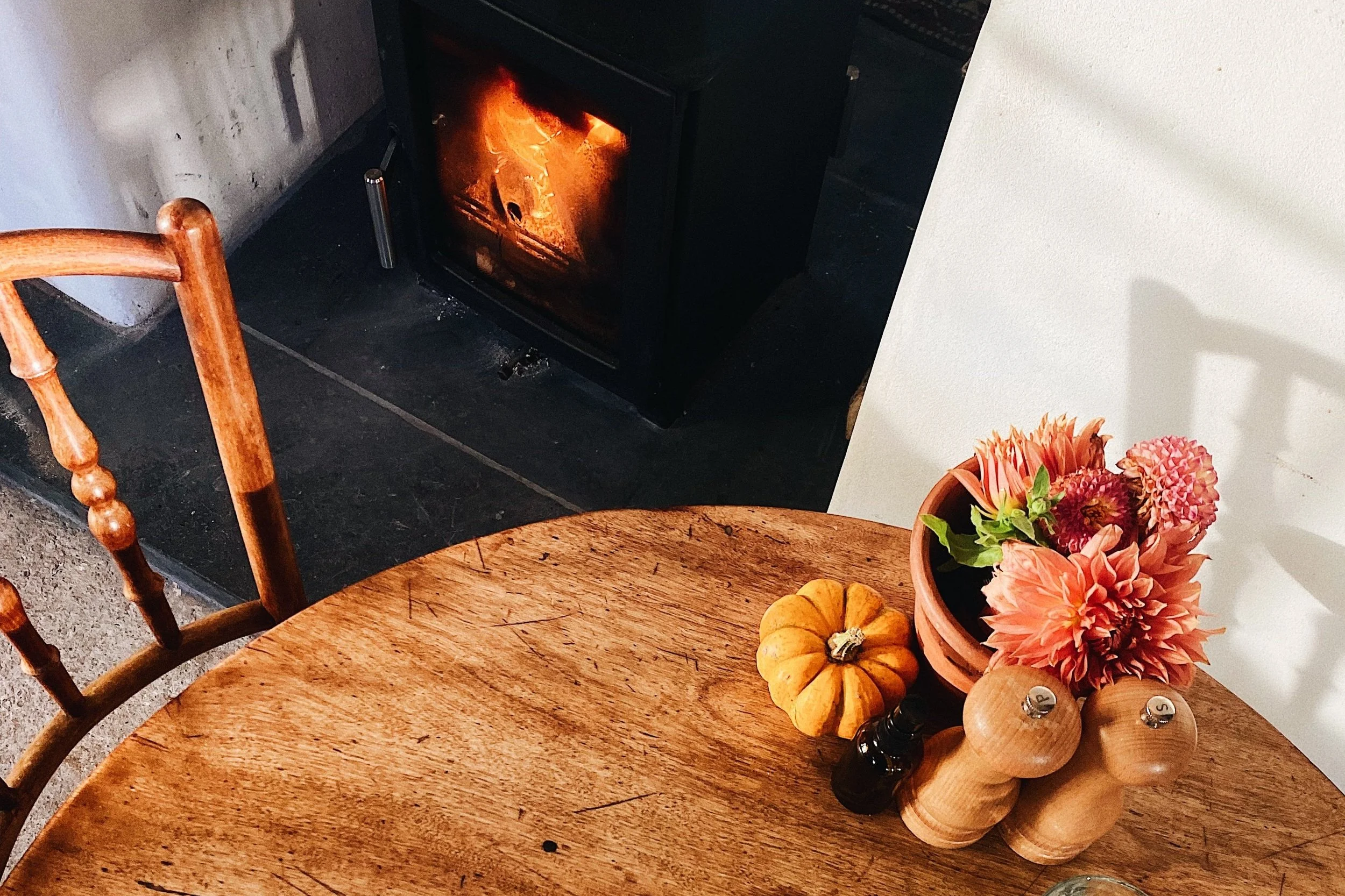A cozy corner featuring a wood stove with visible flames, a round wooden table with a floral arrangement in a terracotta pot, small pumpkins, and wooden salt and pepper shakers.