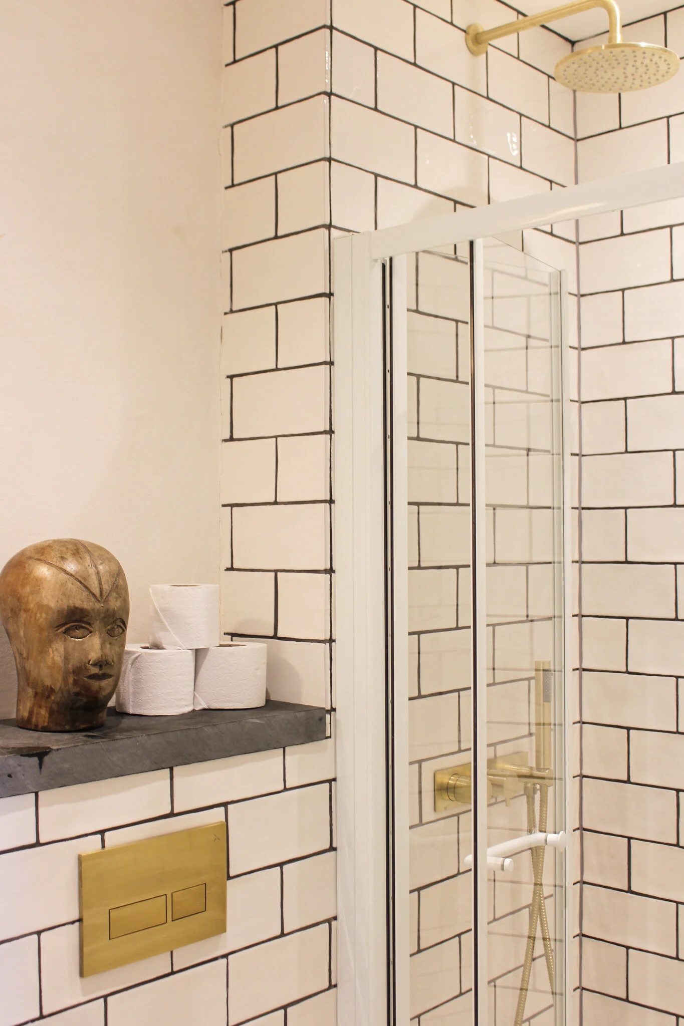 A bathroom shower with white tiled walls outlined in black. The shower has a brass-colored shower head and fixtures. Next to the shower, a shelf holds a wooden tribal mask and rolls of toilet paper, with a white cup behind them.