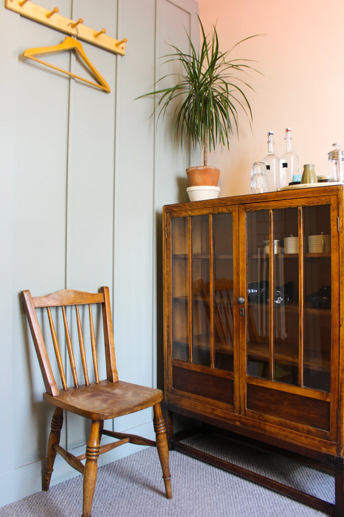 A wooden chair with vertical slats on the backrest in front of a light-colored wall with a mounted coat rack with hooks and a yellow hanger, next to a wooden cabinet with glass doors, a potted plant, and various bottles and glassware on top.