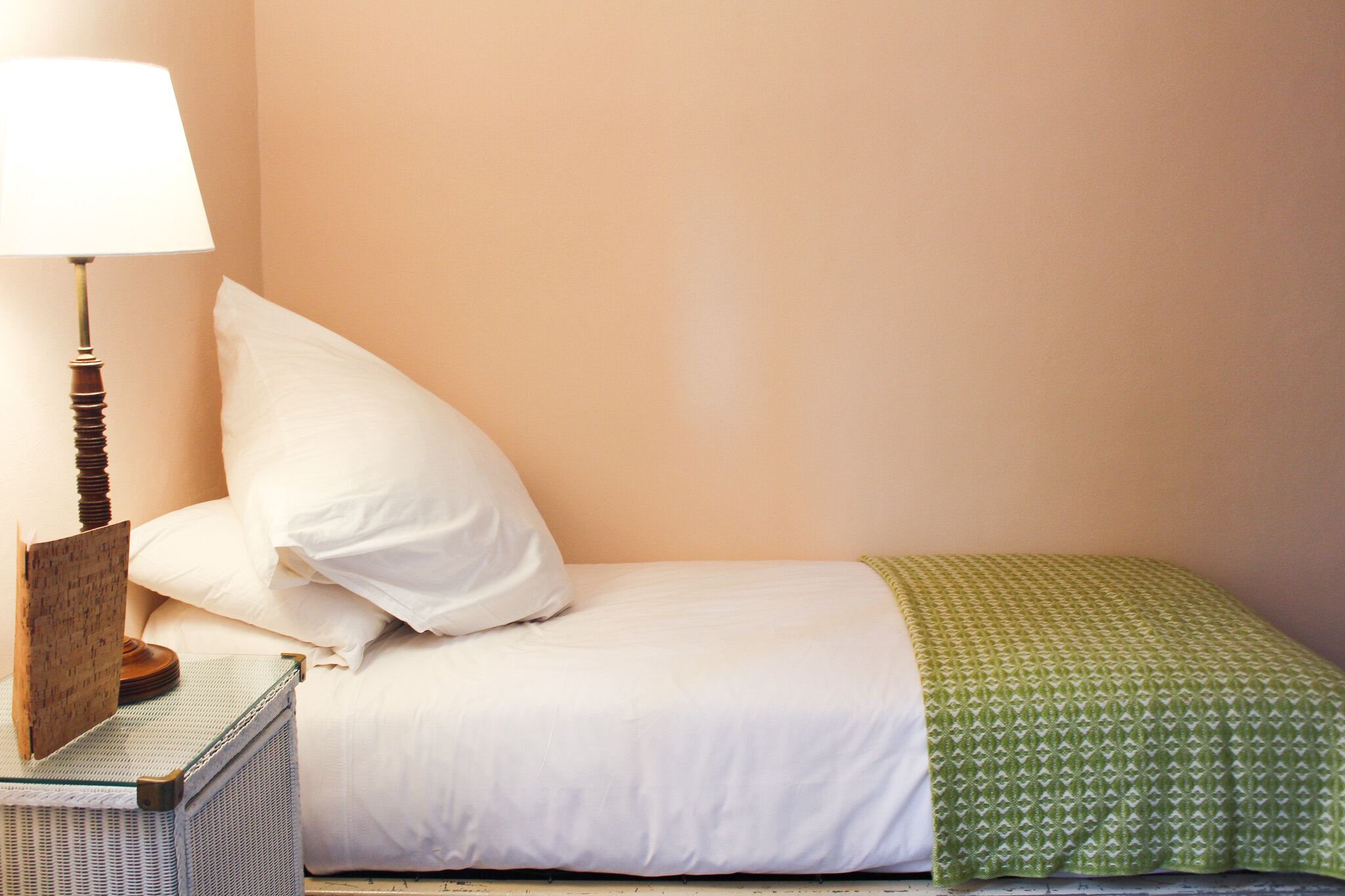 A neatly made hotel bed with white pillows and a green patterned bed runner, next to a nightstand with a lamp and a decorative item, against a beige wall.