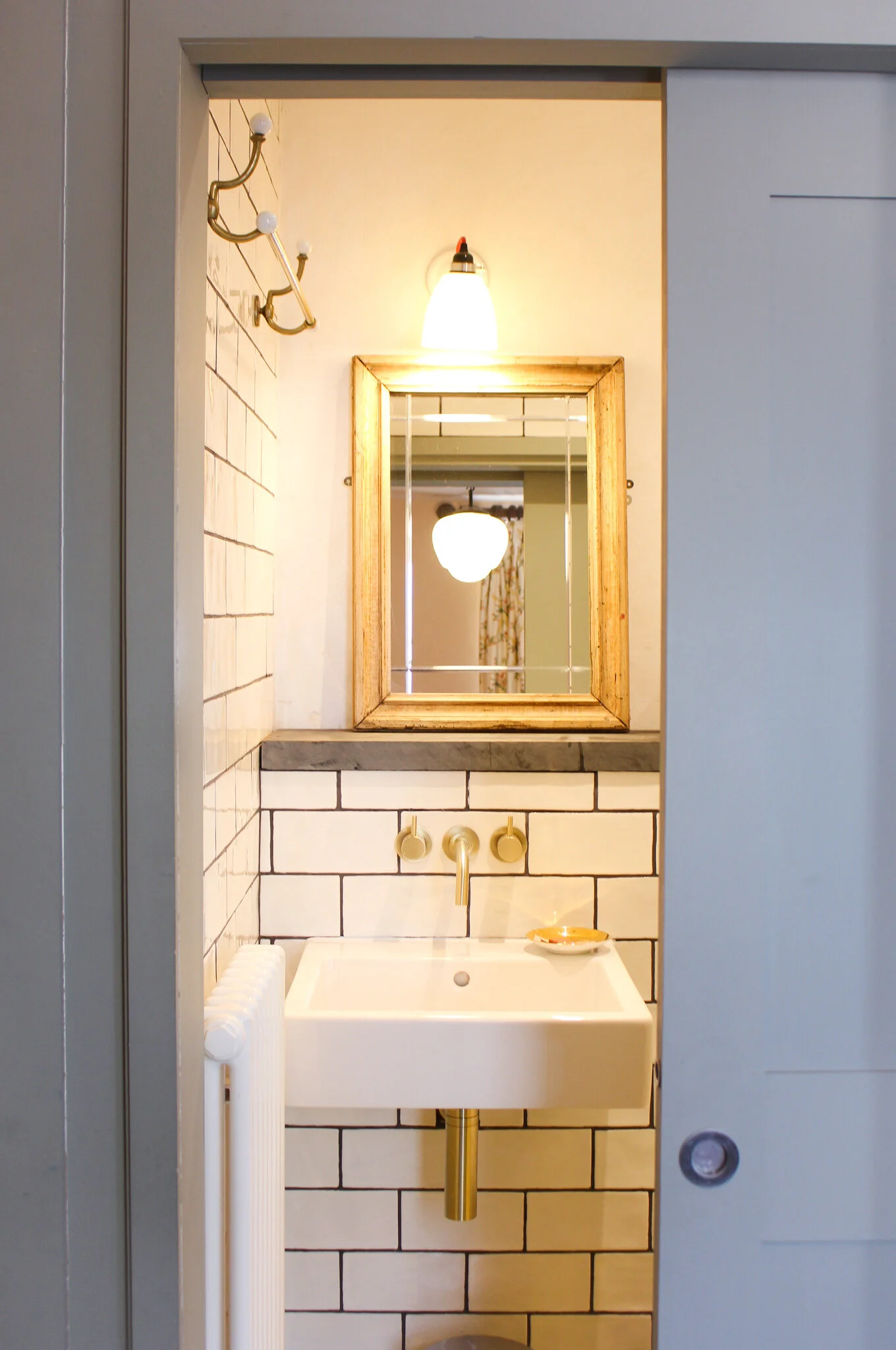 A small vintage bathroom sink with a gold-colored faucet, white tiled walls with black grout lines, a wooden framed mirror, and a wall-mounted light fixture above. A small soap dish sits on the sink.