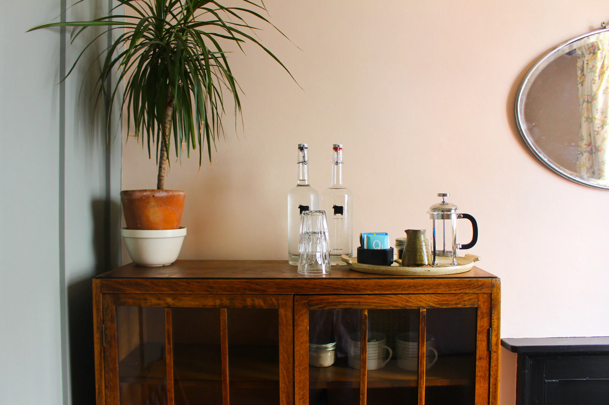 A wooden side table with a potted plant on the left, and various glass bottles, cups, and a teapot on a tray on the right. A round mirror hangs on the wall behind the table.
