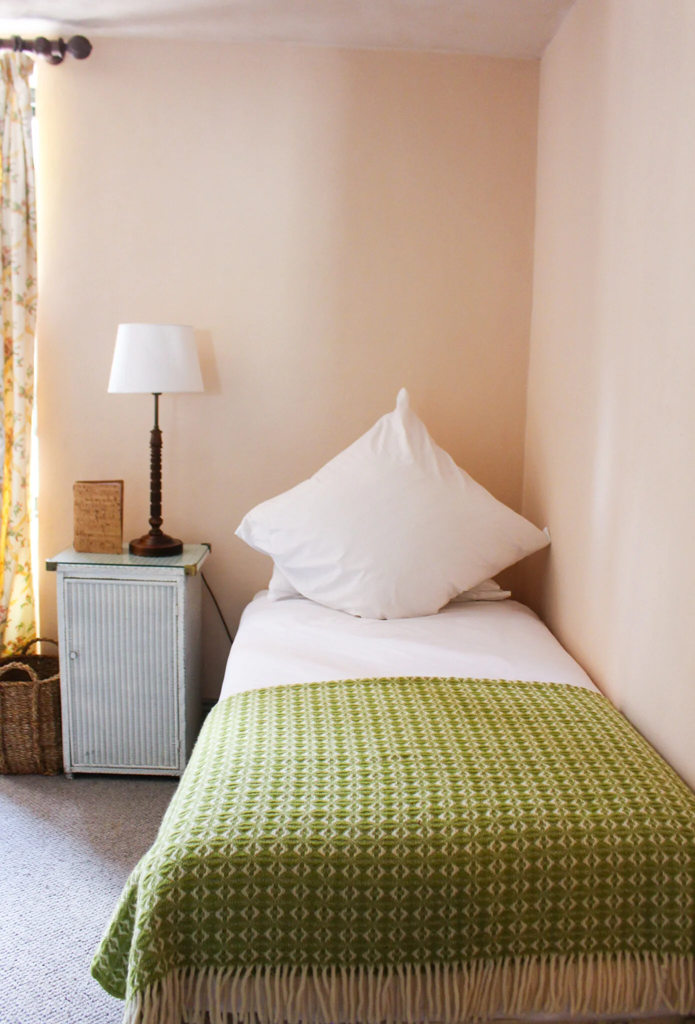 A bedroom with a bed, a green patterned blanket, a white pillow, a nightstand with a lamp and decorative item, beige walls, a window with floral curtains, and a woven basket on the floor.