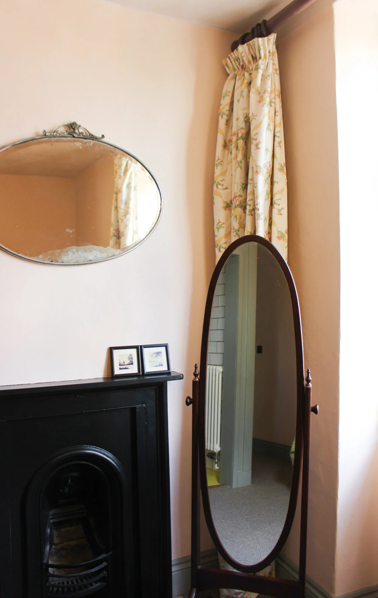 A corner of a room with a black fireplace, an oval decorative mirror on the wall, a standing full-length mirror, a floral curtain, and small framed pictures on the mantel.