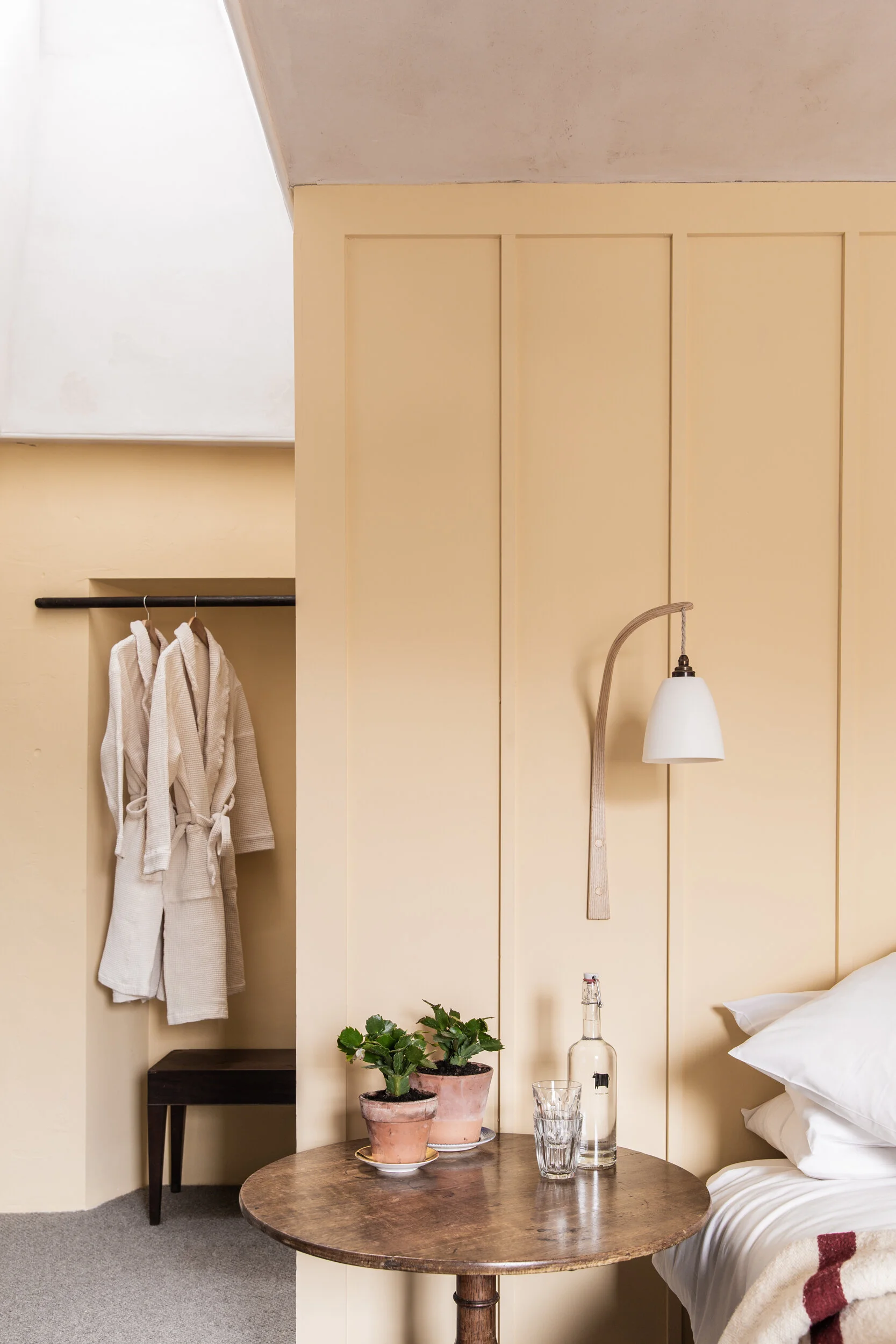 A bedroom corner with a wooden table holding two potted plants, a glass of water, a bottle, and a glass. In the background, there is a bed with white bedding and a beige blanket with a red pattern, a small bedside table, a wall with vertical wood paneling, a hanging white lamp, and a wall shelf with two white robes hanging.