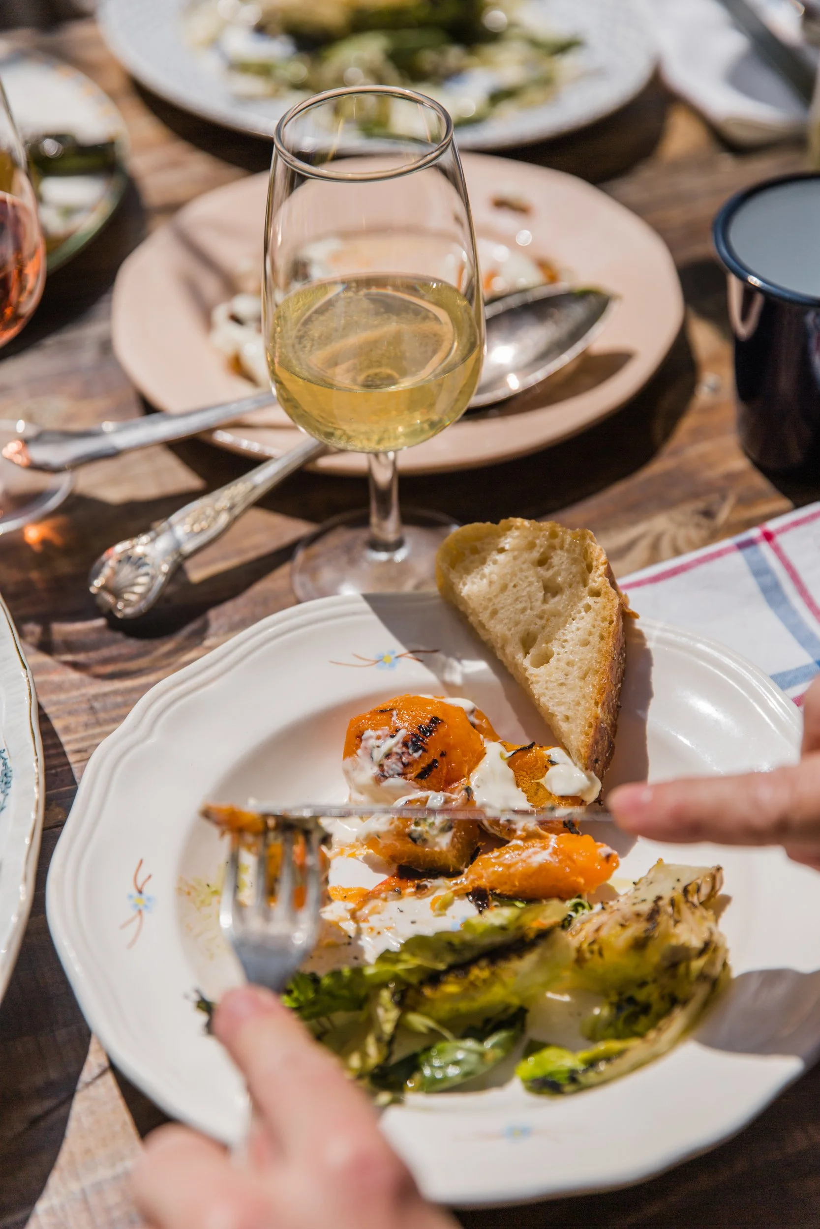 A partially eaten breakfast plate with eggs and vegetables, a piece of bread, and a fork. In the background, a glass of white wine and a partially eaten seafood dish on a pink plate on a wooden table.