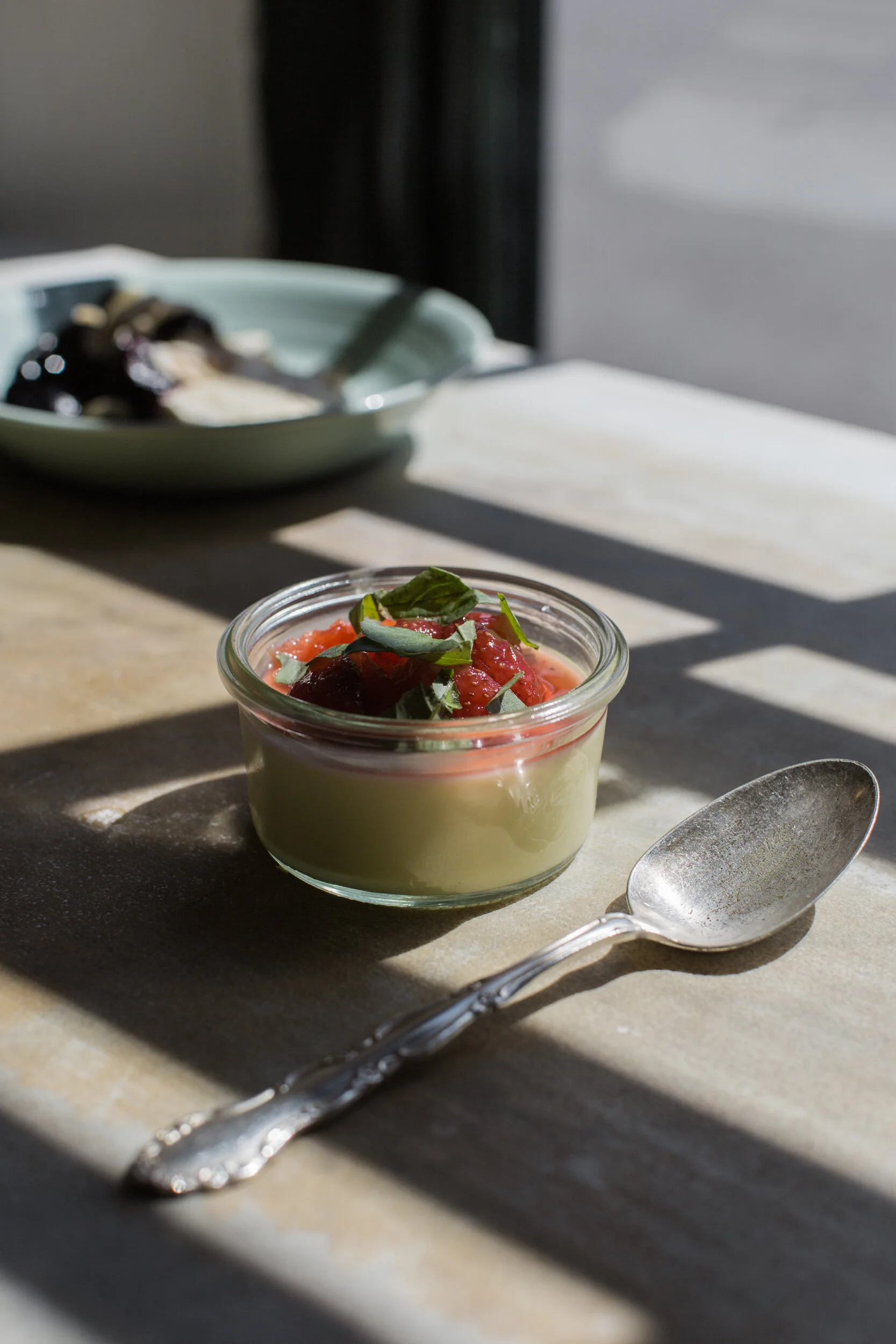 A small glass jar filled with panna cotta topped with mixed berry compote and fresh basil leaves, placed on a beige surface with a silver spoon nearby; blurred plate of berries in the background with sunlight and shadows.