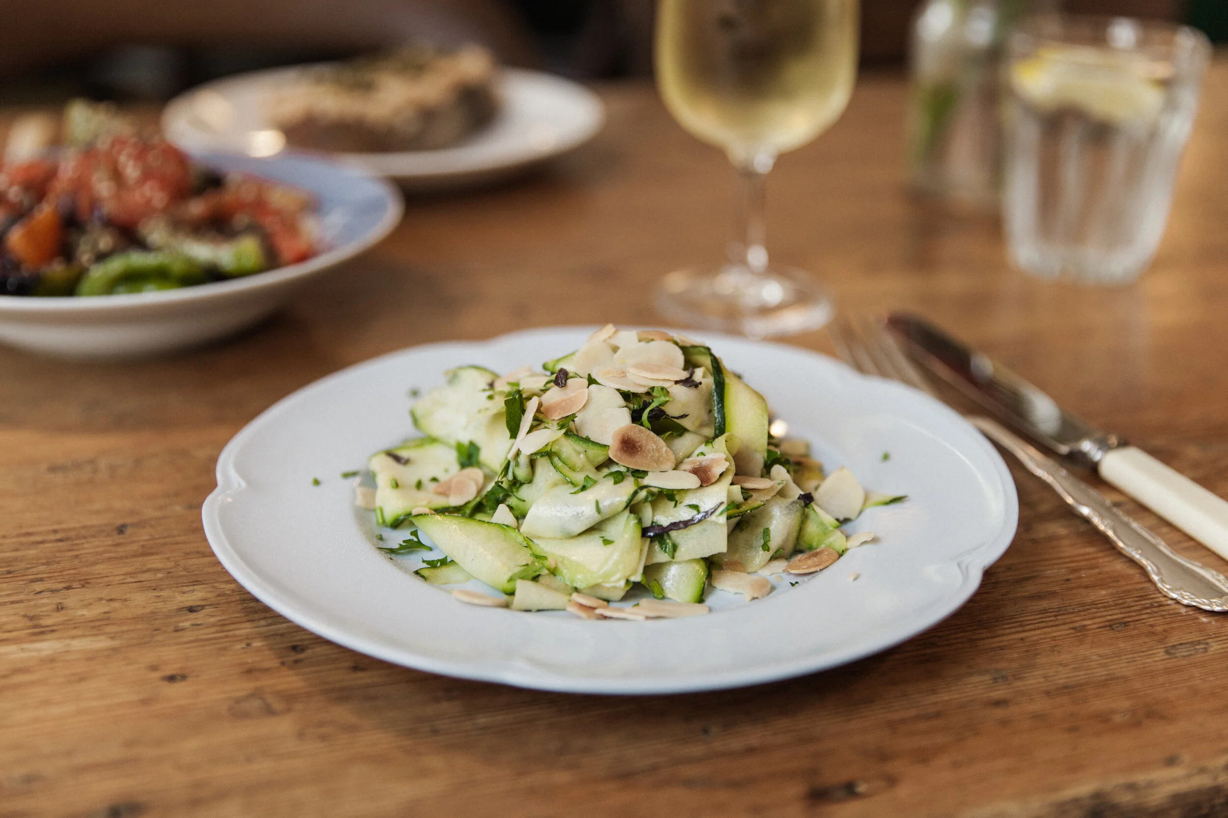 A white plate with cucumber salad topped with sliced almonds and chopped herbs, set on a wooden table with a fork and knife nearby. In the background, there are blurred dishes, a glass of white wine, and a glass of water.