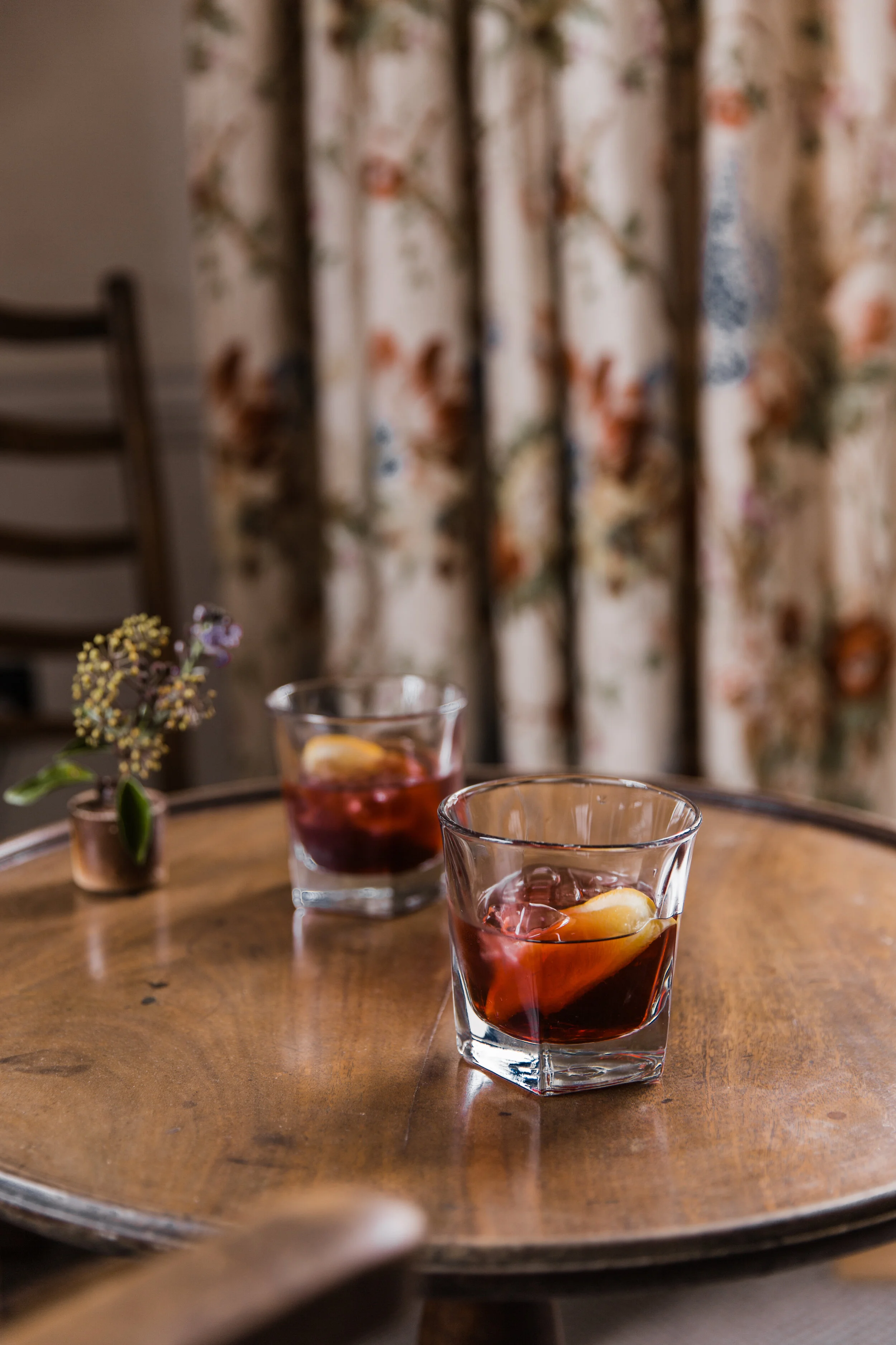 Two glasses of iced tea with lemon slices on a wooden table, with a floral curtain in the background and a small potted plant on the side.
