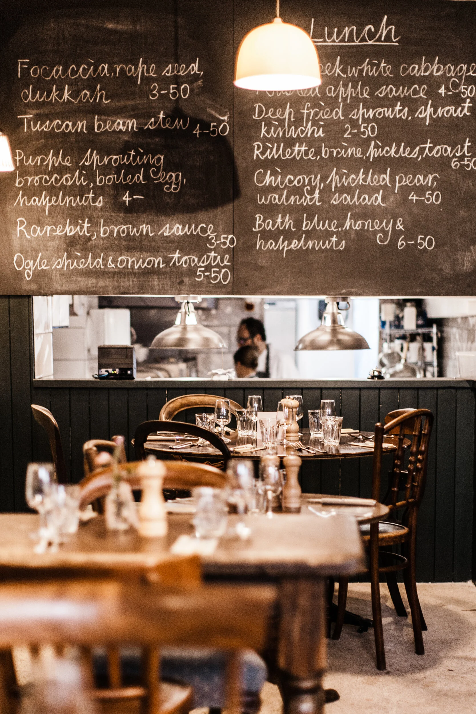 A restaurant interior featuring wooden tables and chairs, a chalkboard menu with handwritten dishes, and warm lighting from pendant lamps, with some patrons visible in the background.