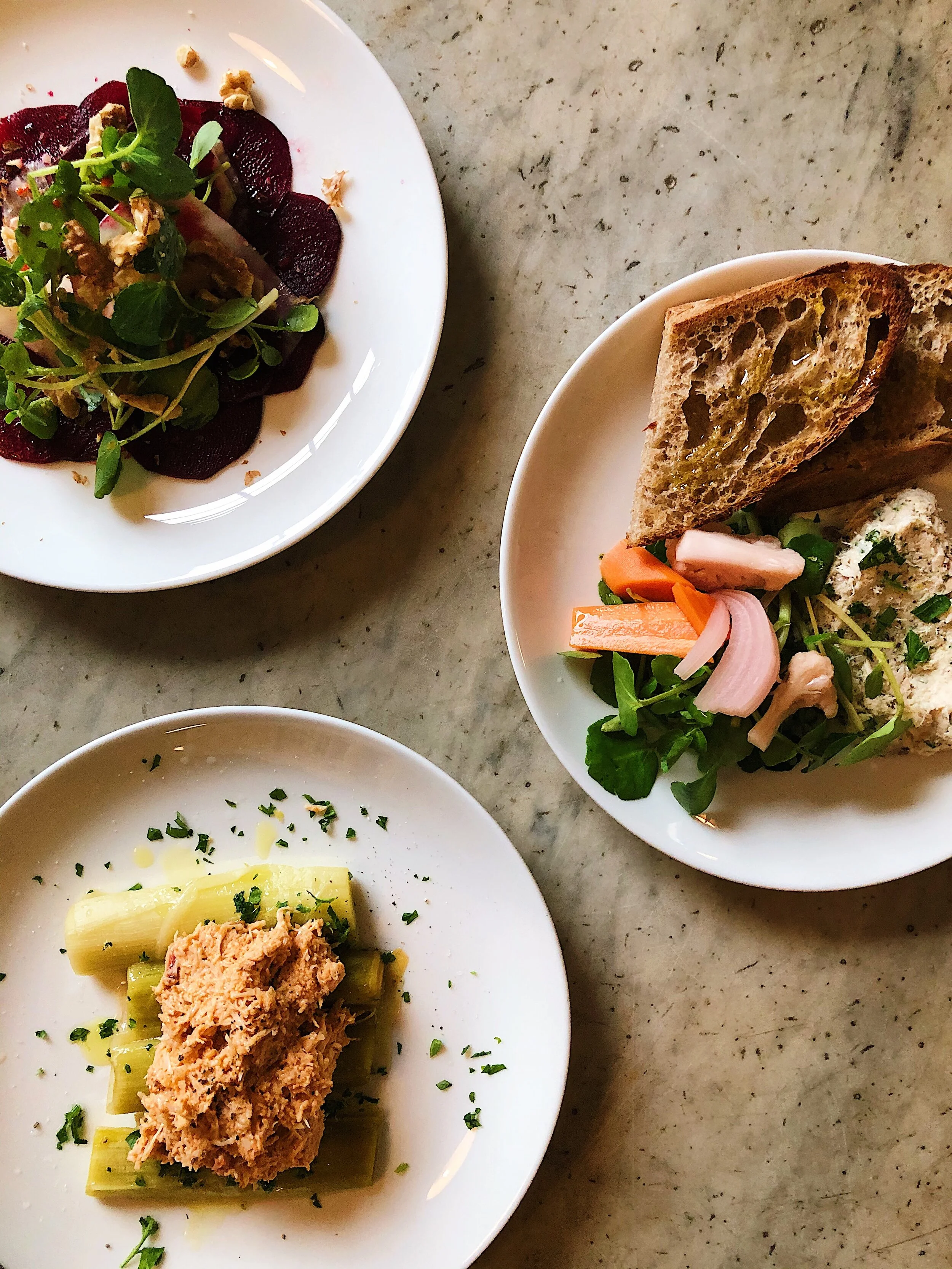 A close-up of a white plate of cooked pasta with a tuna topping, garnished with chopped herbs, and a bowl with a baked slice of bread with olive oil and a side of mixed vegetables including carrots, radishes, and greens. Part of another dish with sli