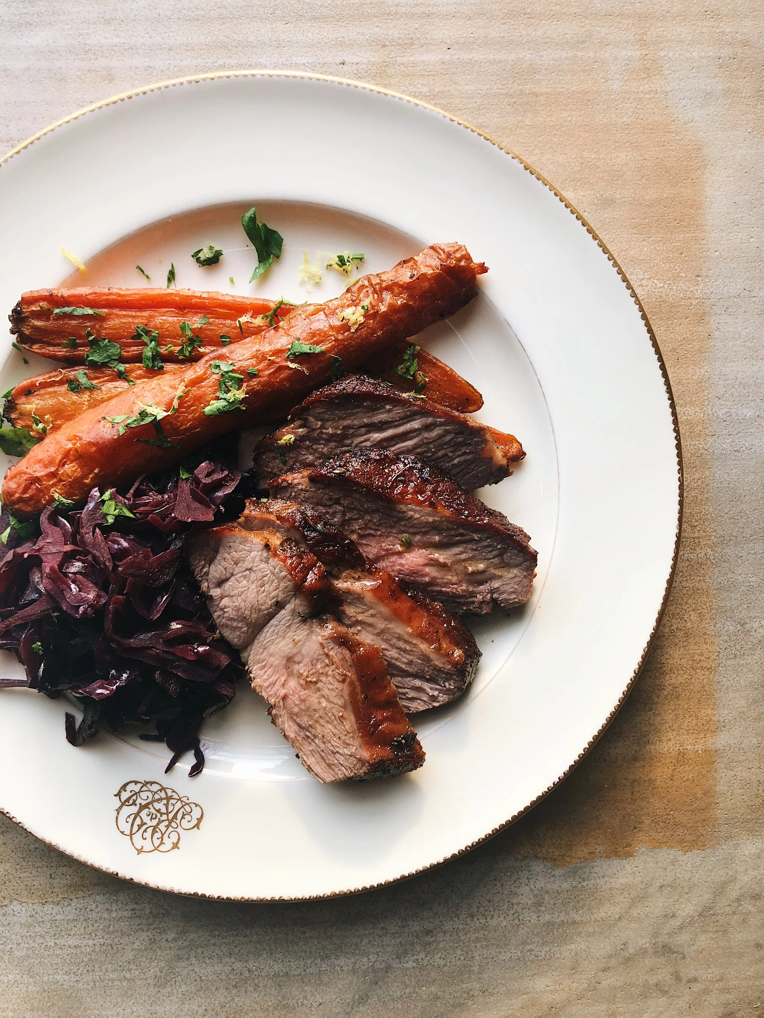 A white decorative plate with a gold rim holding sliced grilled steak, roasted carrots garnished with herbs, and purple shredded cabbage, placed on a beige surface.