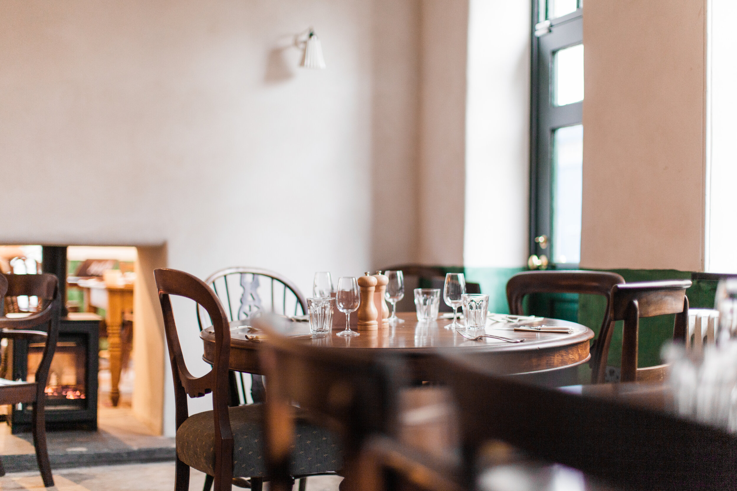 Interior of a restaurant with a wooden table set with glasses, salt and pepper shakers, a napkin, and silverware. There are wooden chairs around the table, large windows letting in natural light, and a plain beige wall with a wall light fixture.