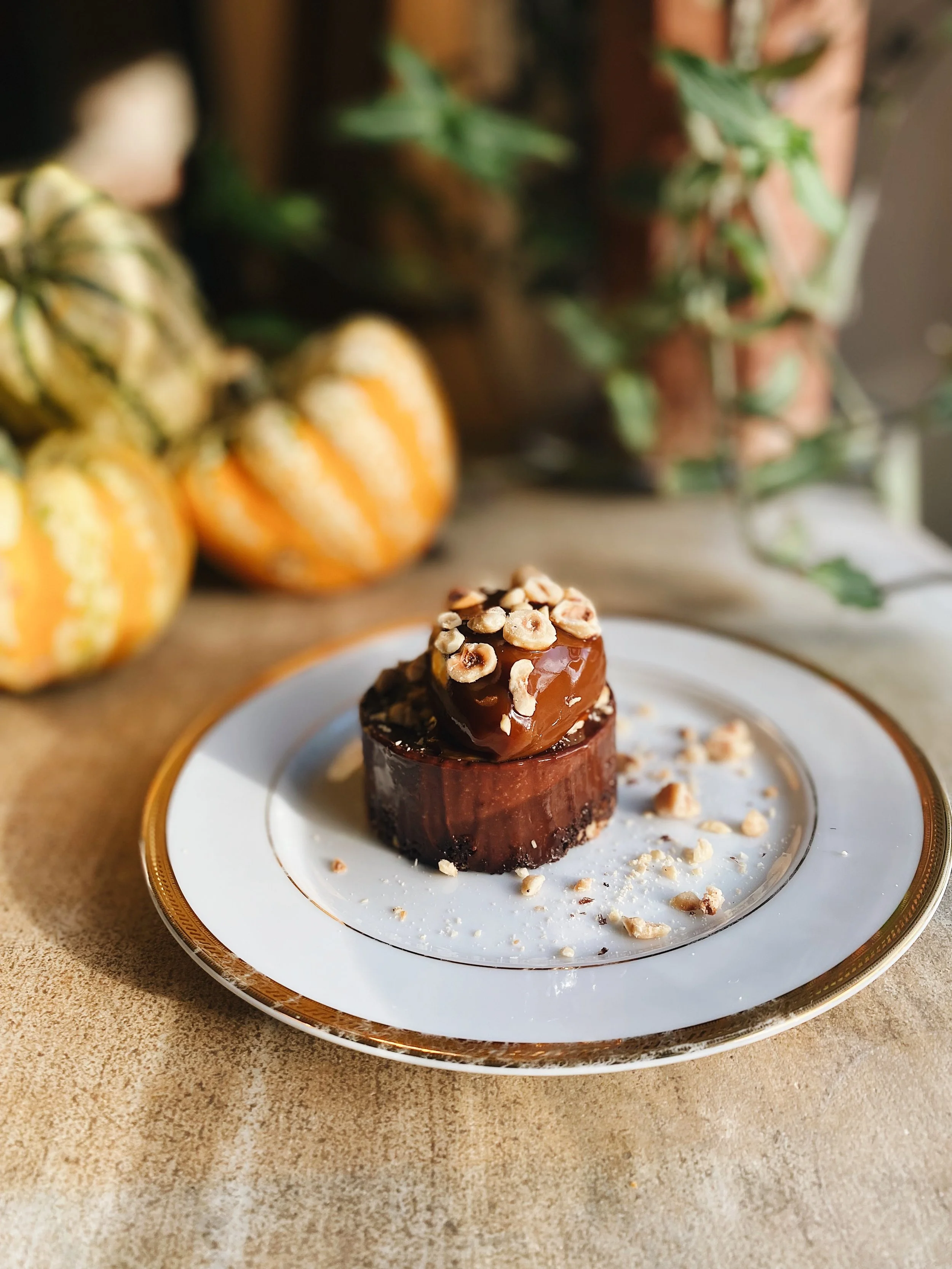 A chocolate dessert topped with chopped nuts on a white plate with a gold rim, placed on a textured beige surface. In the background, there's a blurred display of small decorative pumpkins and green plants.