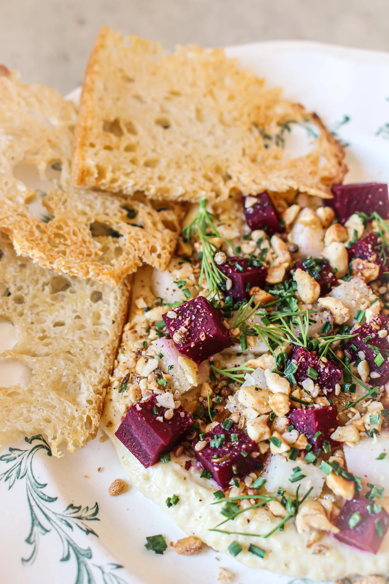 Close-up of a plate with a slice of cheese, beetroot salad topped with chopped nuts and herbs, and crispy bread chips.