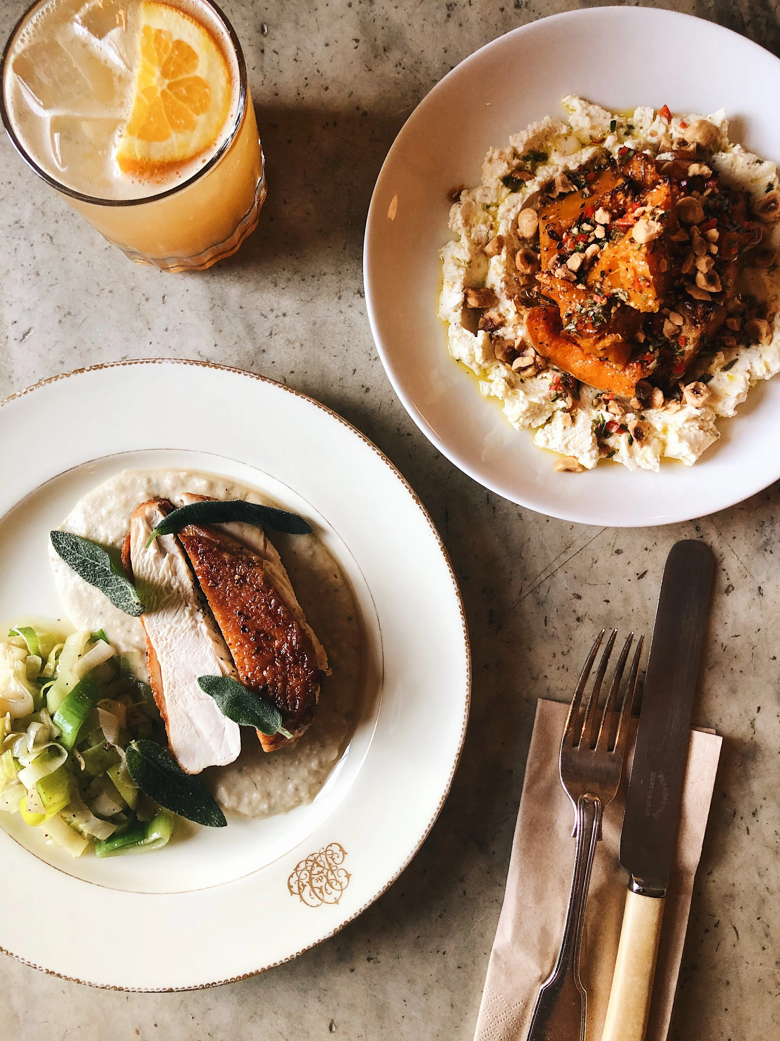 Plate with roasted chicken breast, green vegetables, and mashed potatoes with sage leaves, alongside a glass of orange juice with ice and a slice of orange, and a plate of mashed potatoes with roasted pumpkin and chopped nuts, on a gray speckled tabl